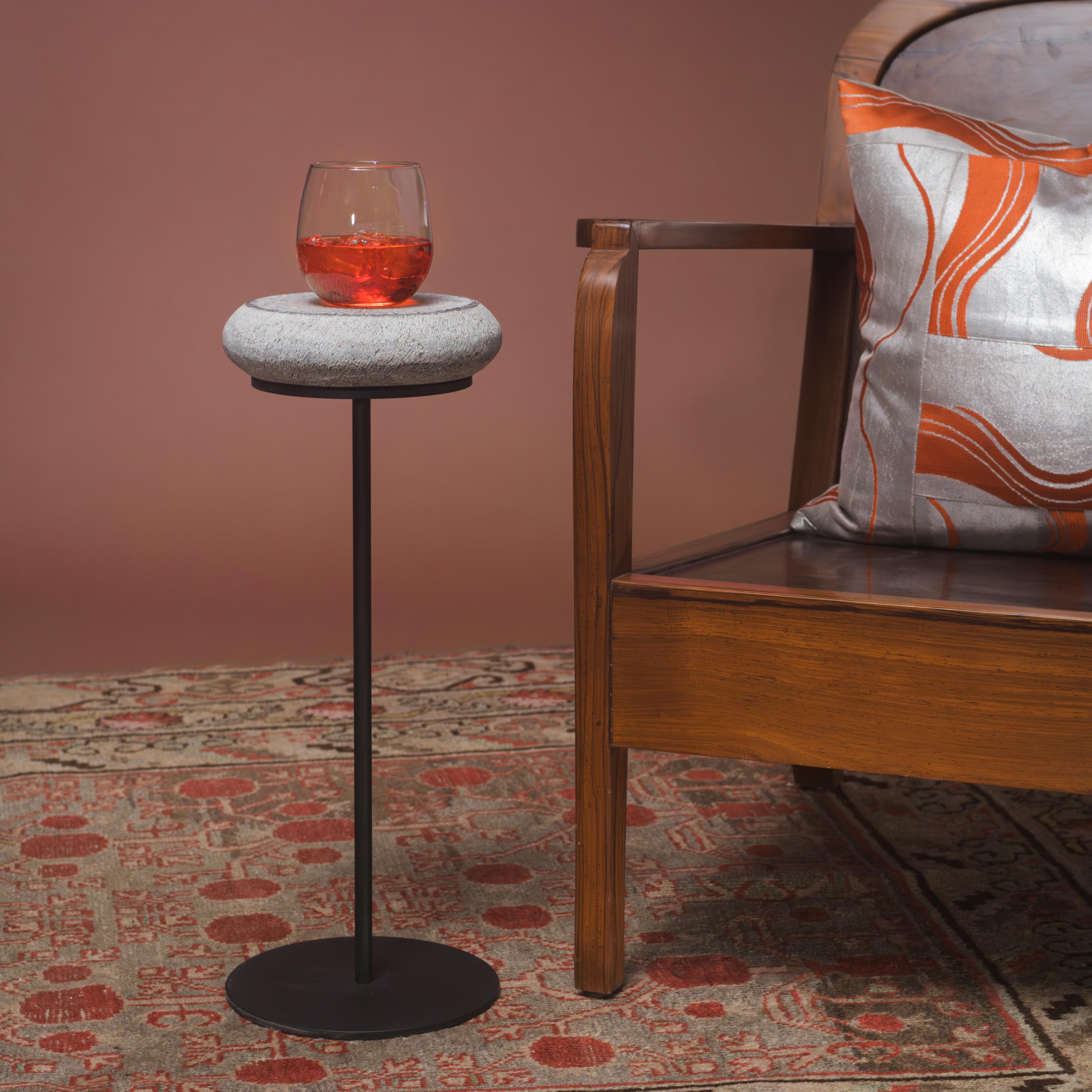 The stone top of this petite side table is carved of solid limestone into the form of a small hand-held drum. The stone drum has a simple, round form with curved sides and a textured surface. Placed atop a matte steel pedestal base, this small