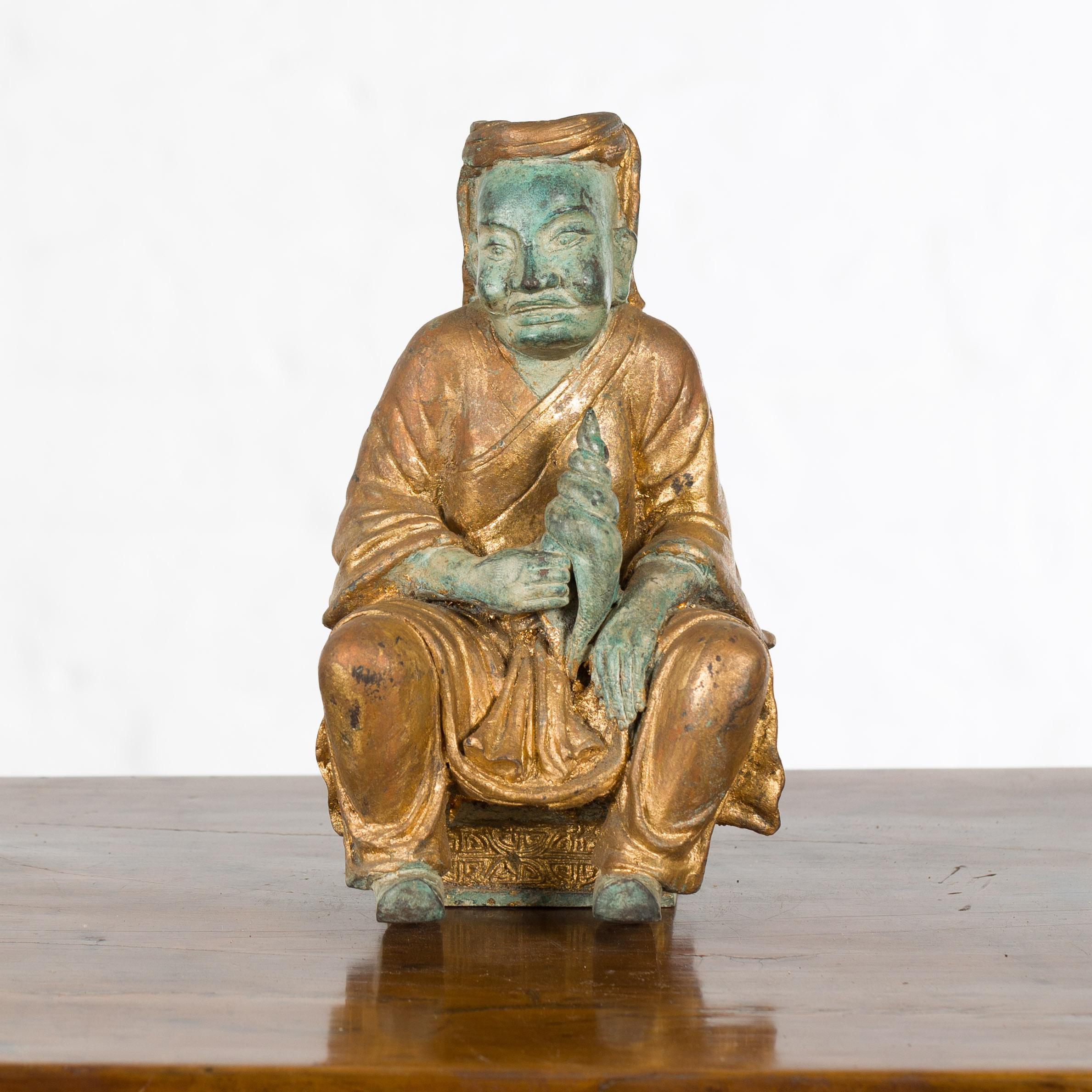 A small Thai vintage seated monk holding a shell from the mid 20th century with verde and gilt bronze finish. Created in Thailand during the mid century period, this petite bronze sculpture features a seated monk holding a conch shell in his right