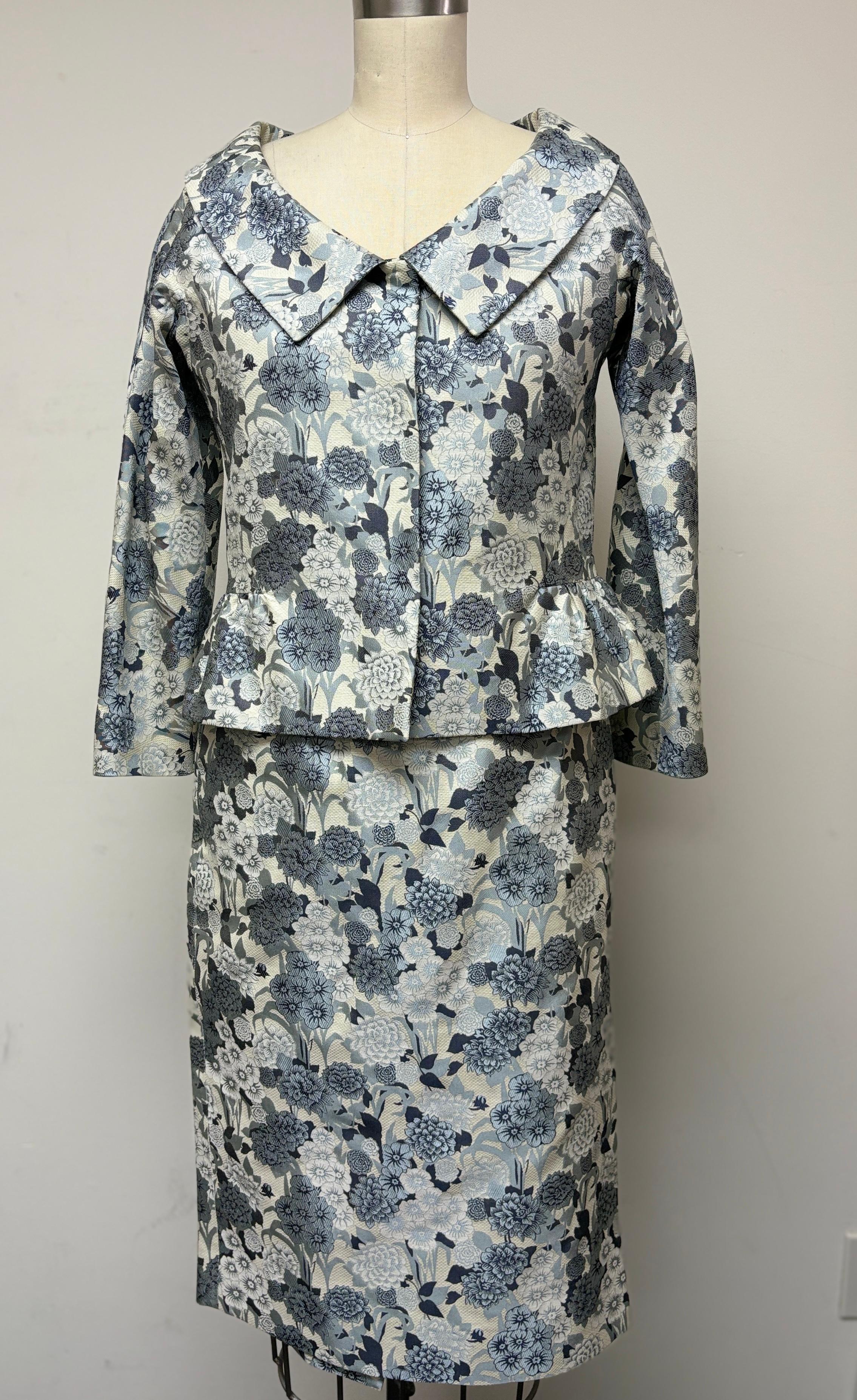 A stunning floral skirt suit featuring various pewter, blue, black, and white flowers all over. This classic look immerses you into a spring-like garden party. This dazzling jacket features a snap front, a beautiful round collar, and a gathered
