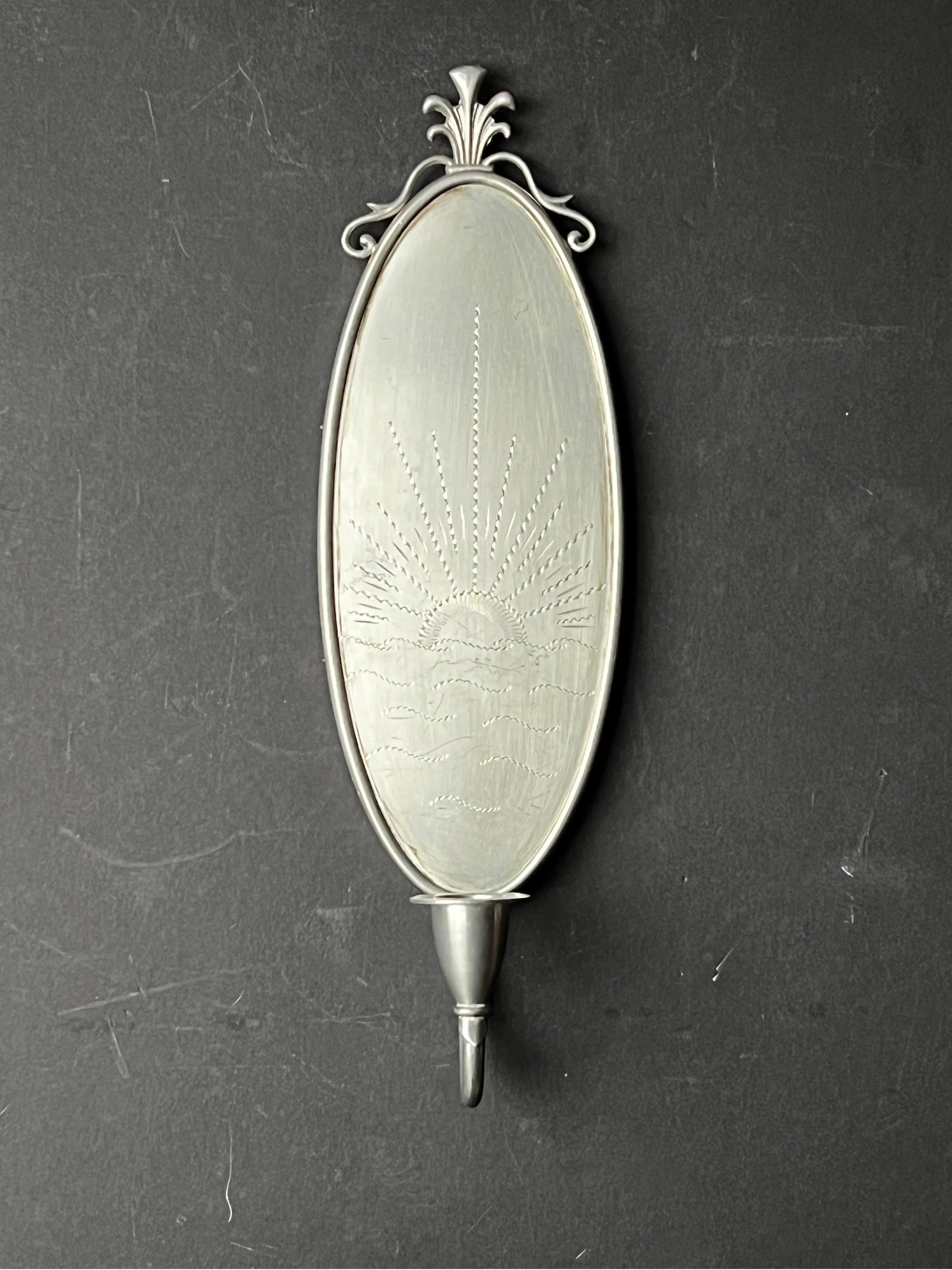 An oval candle sconce in silver-coloured metal - mostly likely pewter. The oval panel is engraved with a simple design of a sunrise (or sunset) over the sea. The wooden back has a single hanging point which is stamped 
