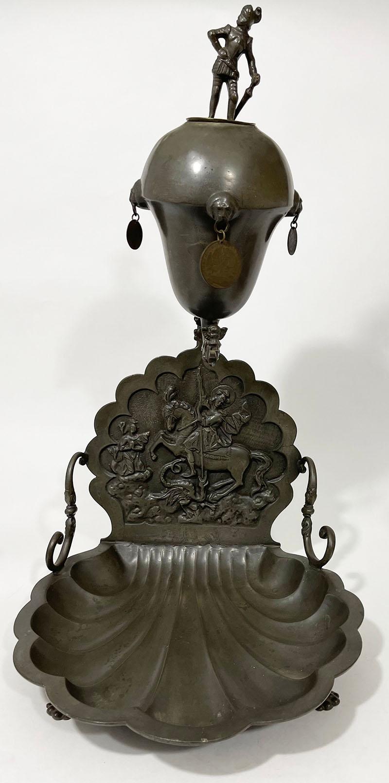 Pewter fountain and basin with a scene of Saint George and the dragon

A pewter shell-shaped basin, raised on two claw feet with a scalloped rim and a straight, upright backplate with a relief scene of Saint George and the dragon, supported by two