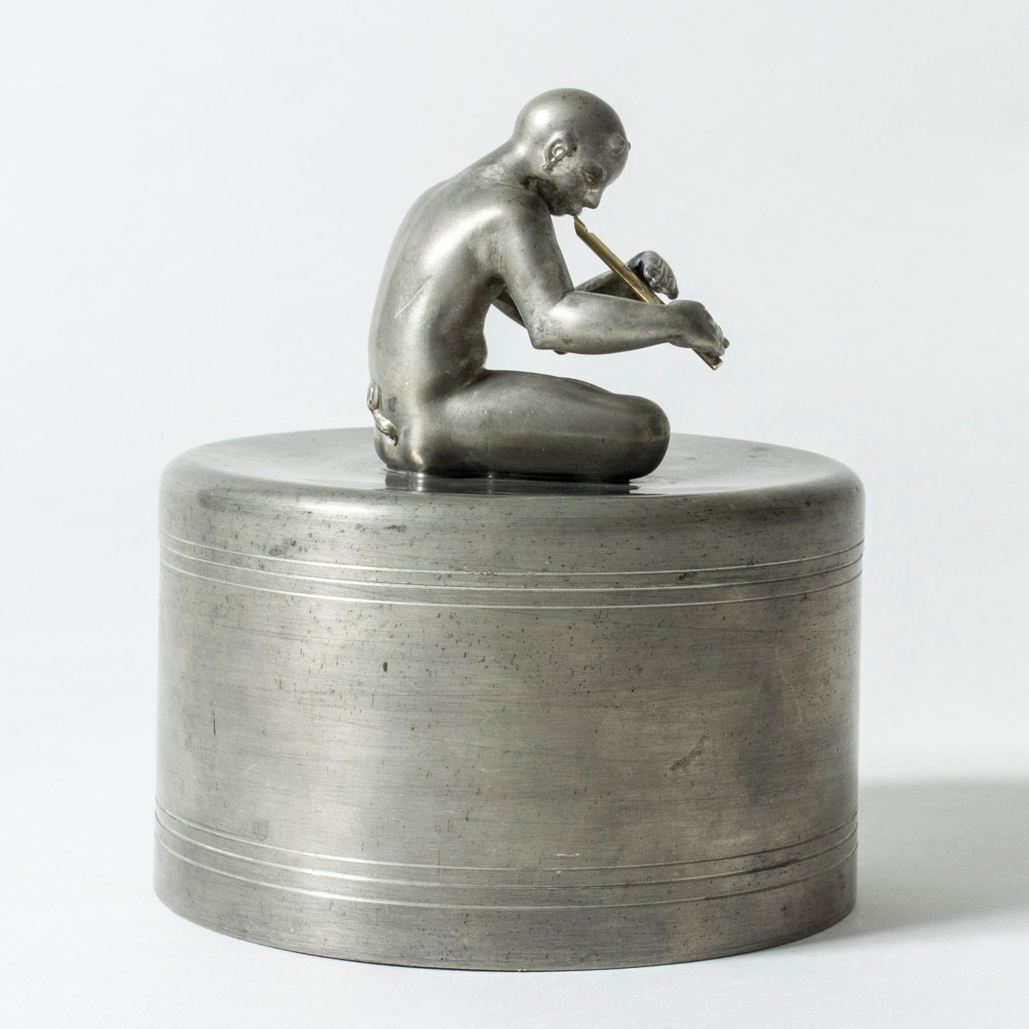 Étonnante jarre en étain de Nils Fougstedt, ornée d'une figurine de Pan jouant de la flûte, les jambes croisées. La petite flûte est fabriquée en laiton. Figurine magnifiquement sculptée avec une attention particulière aux détails et une pose