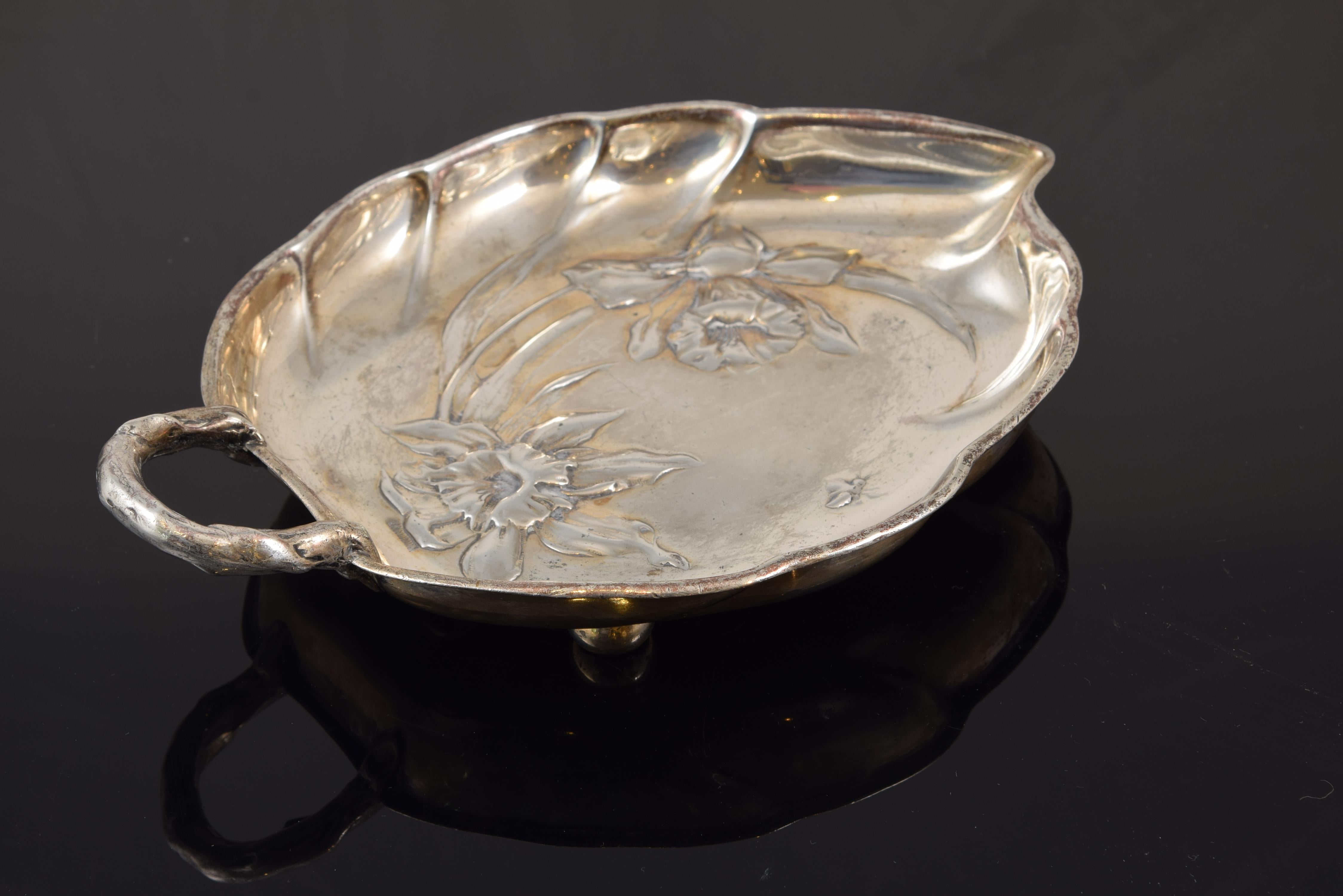 Bandeja de estilo modernista (Jugendstil Art Nouveau). Peltre. Kayserzinn, Alemania, hacia 1900.
De marca.
Bandeja elevada de peltre plateado en forma de hoja sobre patas esféricas, con asa y decoración temática floral ligeramente repujada.