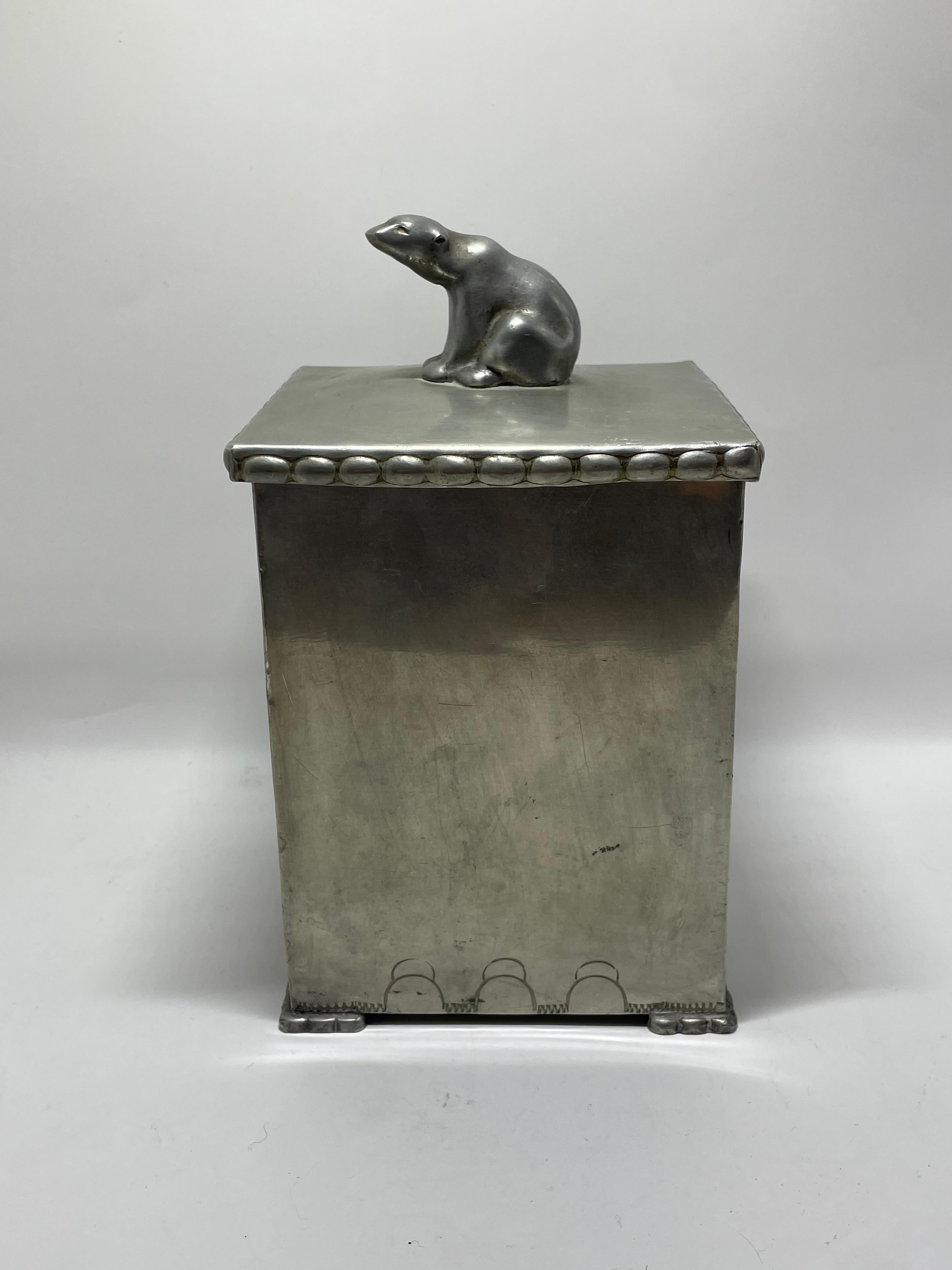 Este recipiente cuadrado presenta una tapa abovedada adornada con una figura de oso polar finamente modelada como remate, un detalle distintivo que aporta carácter y fantasía al diseño. El perímetro de la tapa está decorado con un borde de cuentas,