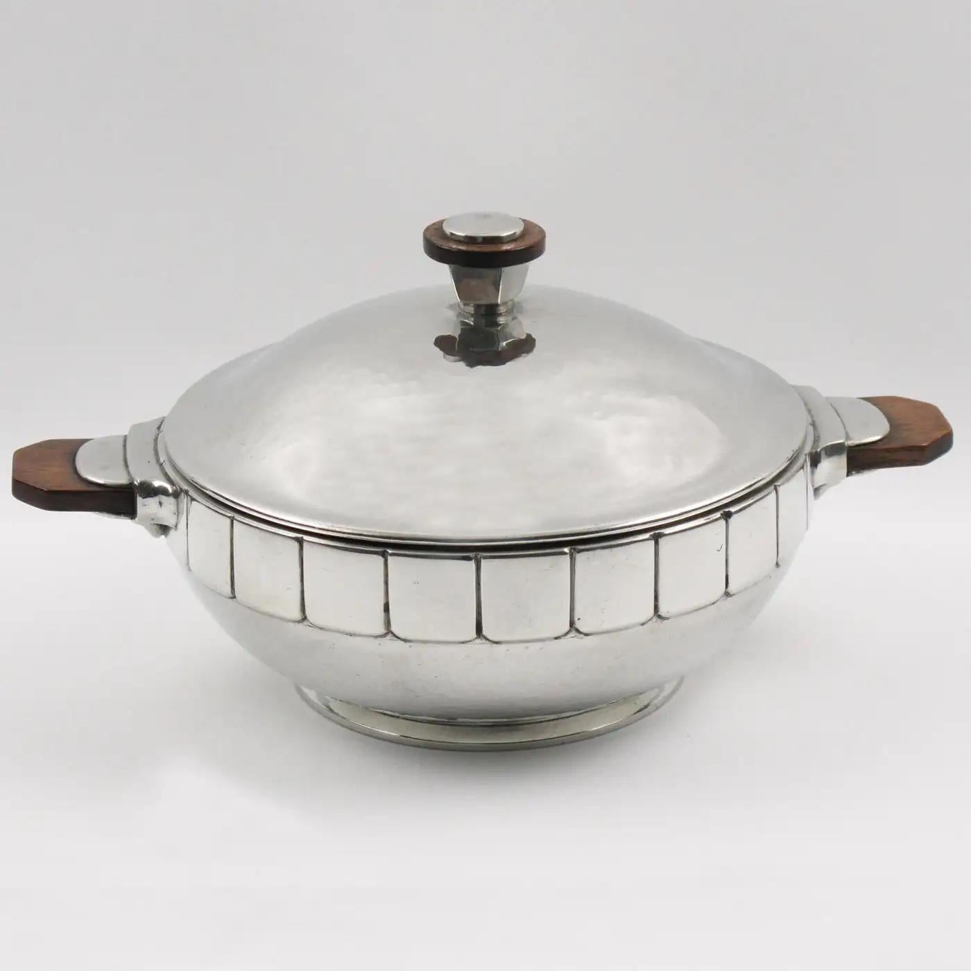 This lovely embossed polished pewter tureen was designed and manufactured by H. J. Swiss Pewter. The modernist shape has a detailed embossed design and solid wood handles. The covered dish is marked underside with Eagle/Phoenix head emblem and H.J.