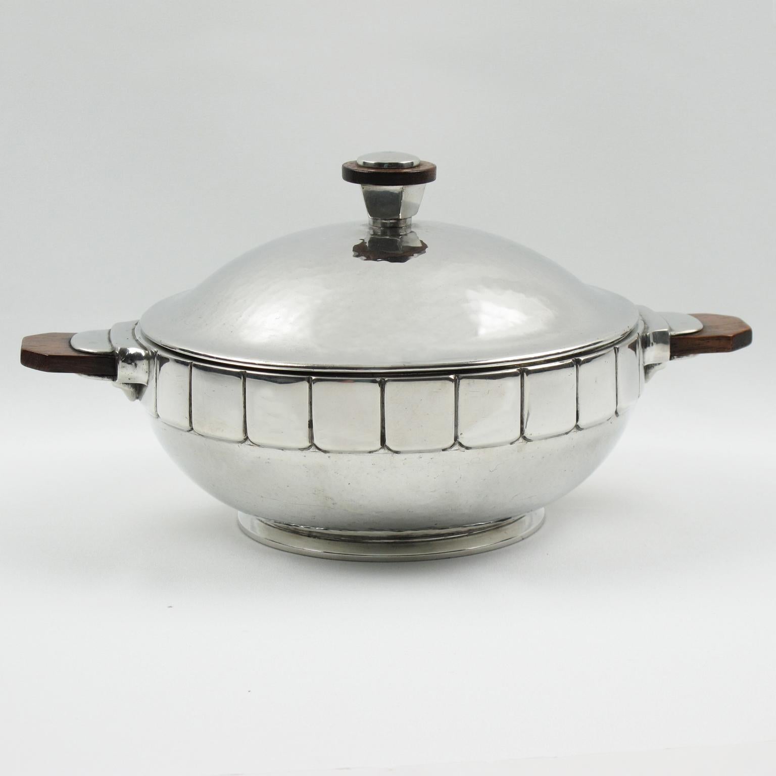 Esta preciosa sopera de peltre pulido en relieve fue diseñada y fabricada por H. J. Swiss Pewter. La forma modernista tiene un detallado diseño en relieve y asas de madera maciza. El plato cubierto lleva en la parte inferior el emblema de la cabeza