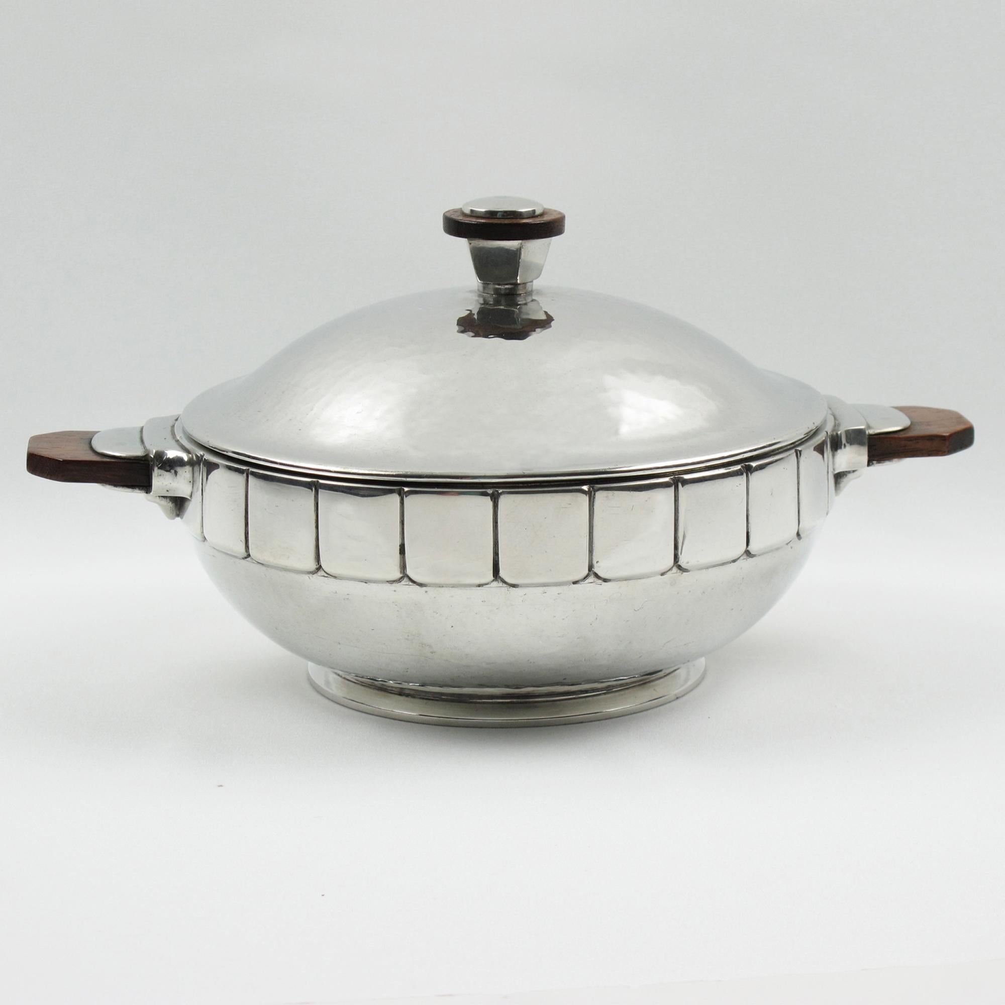 This stunning, embossed, and polished pewter tureen was designed and crafted by H.J. Swiss Pewter. The modernist shape has a detailed embossed design and solid wood handles. The covered dish is marked on the underside with an Eagle/Phoenix head