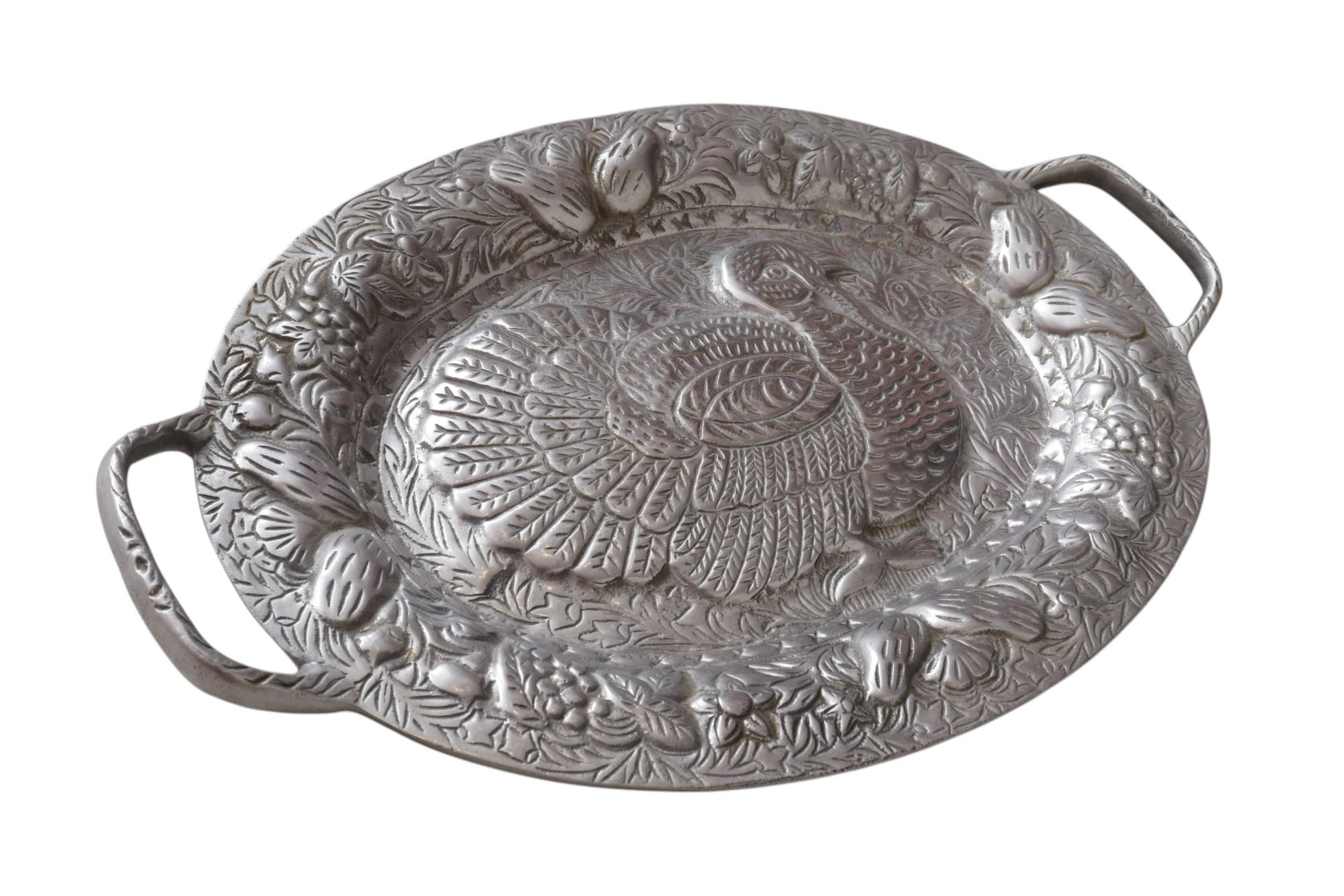 An oval serving platter in pewter, embossed with a turkey surrounded with foliage, fruit and berries, in high relief. The platter has a handle on each side.
