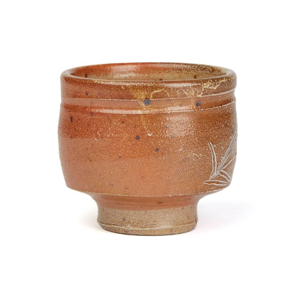 A fine vintage British studio pottery yunomi (teacup) by renowned potter Phil Rogers (b. 1951) and made in Powys, Wales. The stoneware yunomi is of rounded cylindrical shape standing on a narrow rounded foot and is decorated with incised grasses