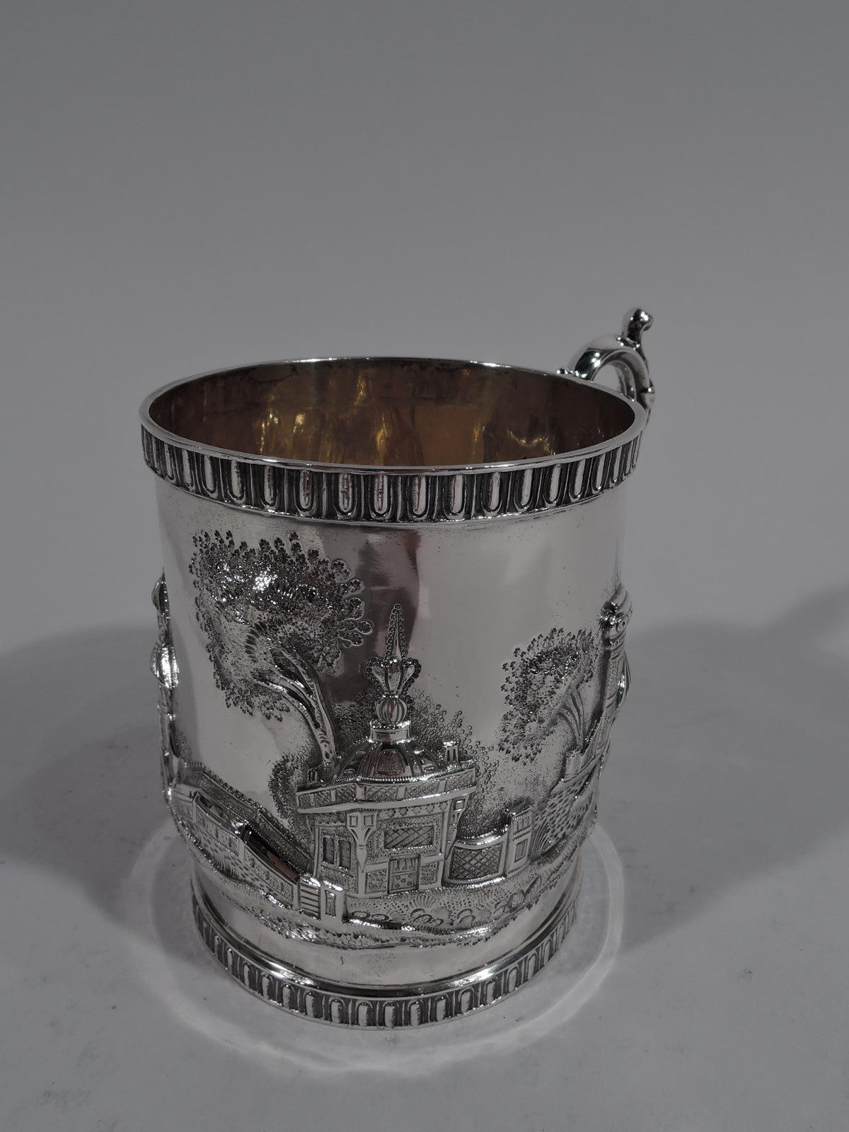 Architectural coin silver baby cup, mid-19th century. Straight sides with leaf-capped and mounted s-scroll handle. Repousse fantasy buildings including turret, pagoda, pavilion, and picturesque bridge. Stylized egg-and-dart rims. Gilt-washed
