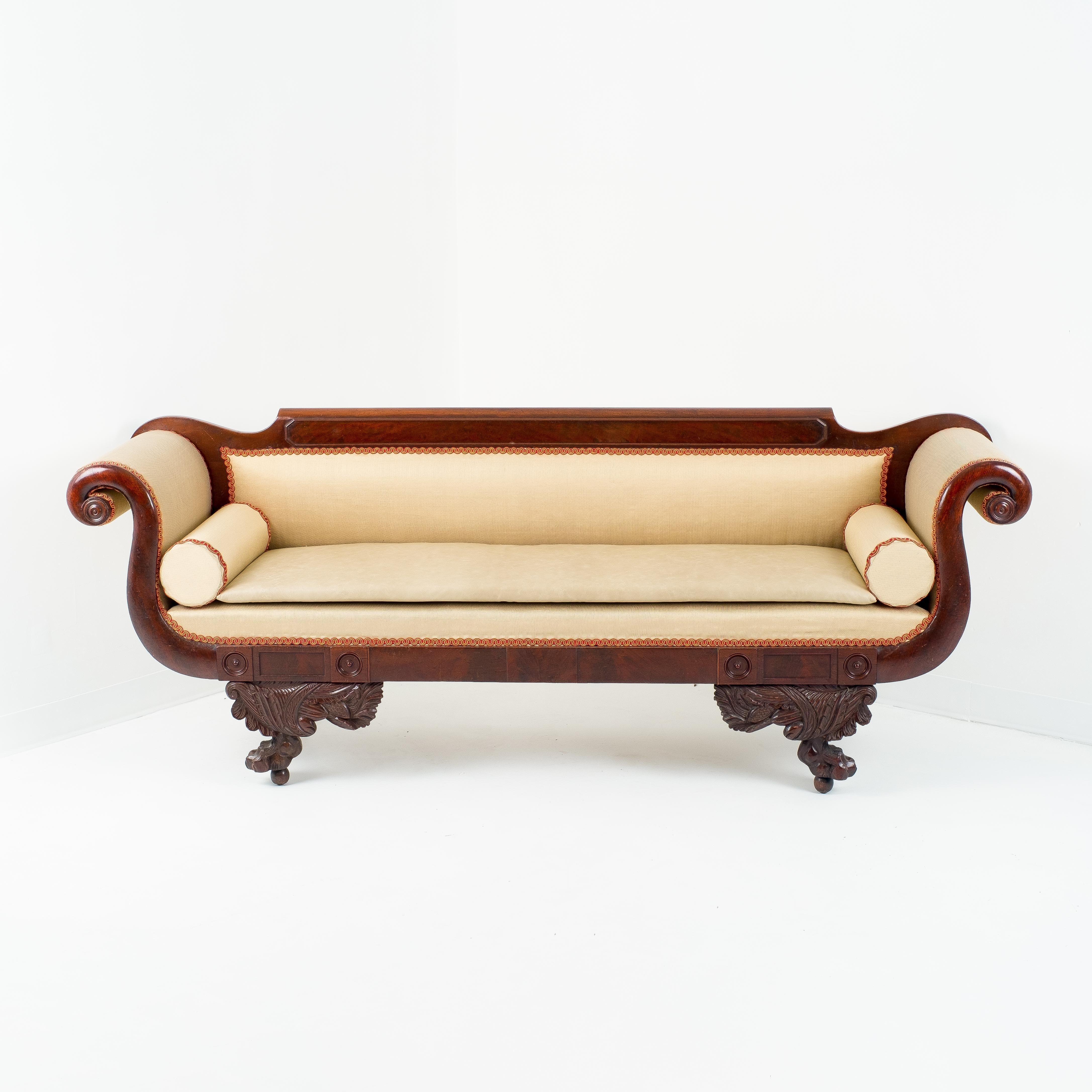 American Neoclassic sofa with mahogany frame upholstered in silk with contrasting gimp. The crest rail has a panel of branch crotch mahogany book matched across the length of the sofa back. The scrolled arms, lion’s paw legs with cornucopia carved