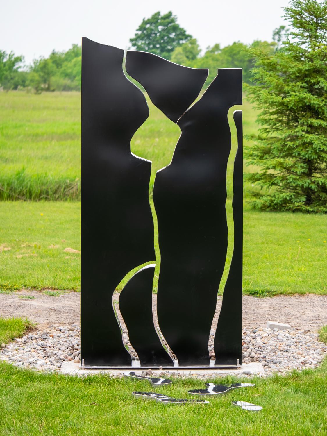 A rectangular, steel outdoor sculpture in black is divided in parts, like a puzzle, the edges polished and reflective. This sculpture Water Writing V3, titled in French as Aquagraphie Variation No 2, is an eight foot work by Quebec artist Philippe
