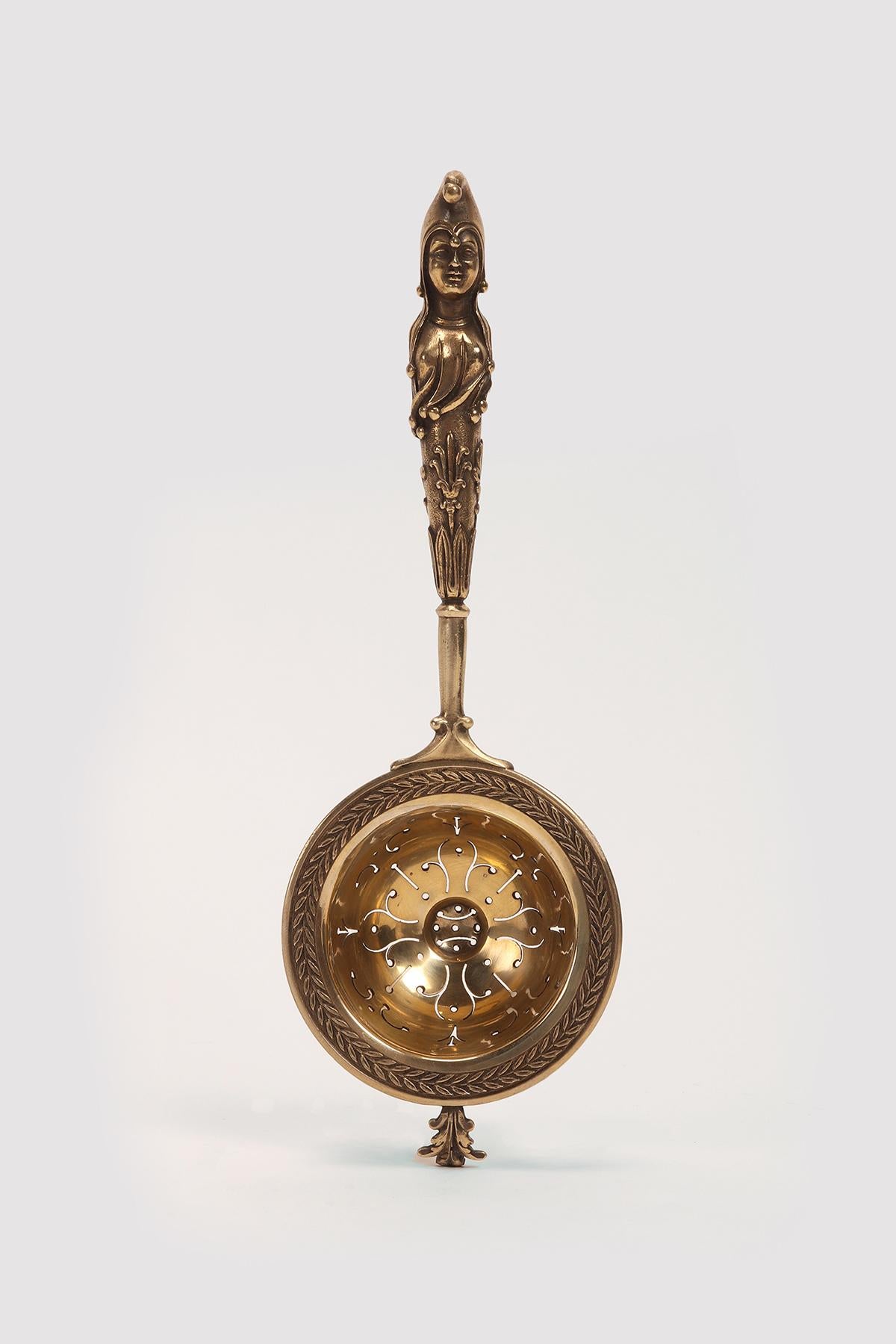 Colino da tè con manico lungo, in argento dorato, con tazza liscia e perforata, peduncolo fitiforme e alloro inciso per decorare il bordo estroflesso. Maniglia con attacco liscio che poi diventa alloro e altri racemi. In alto, l'erma di una donna