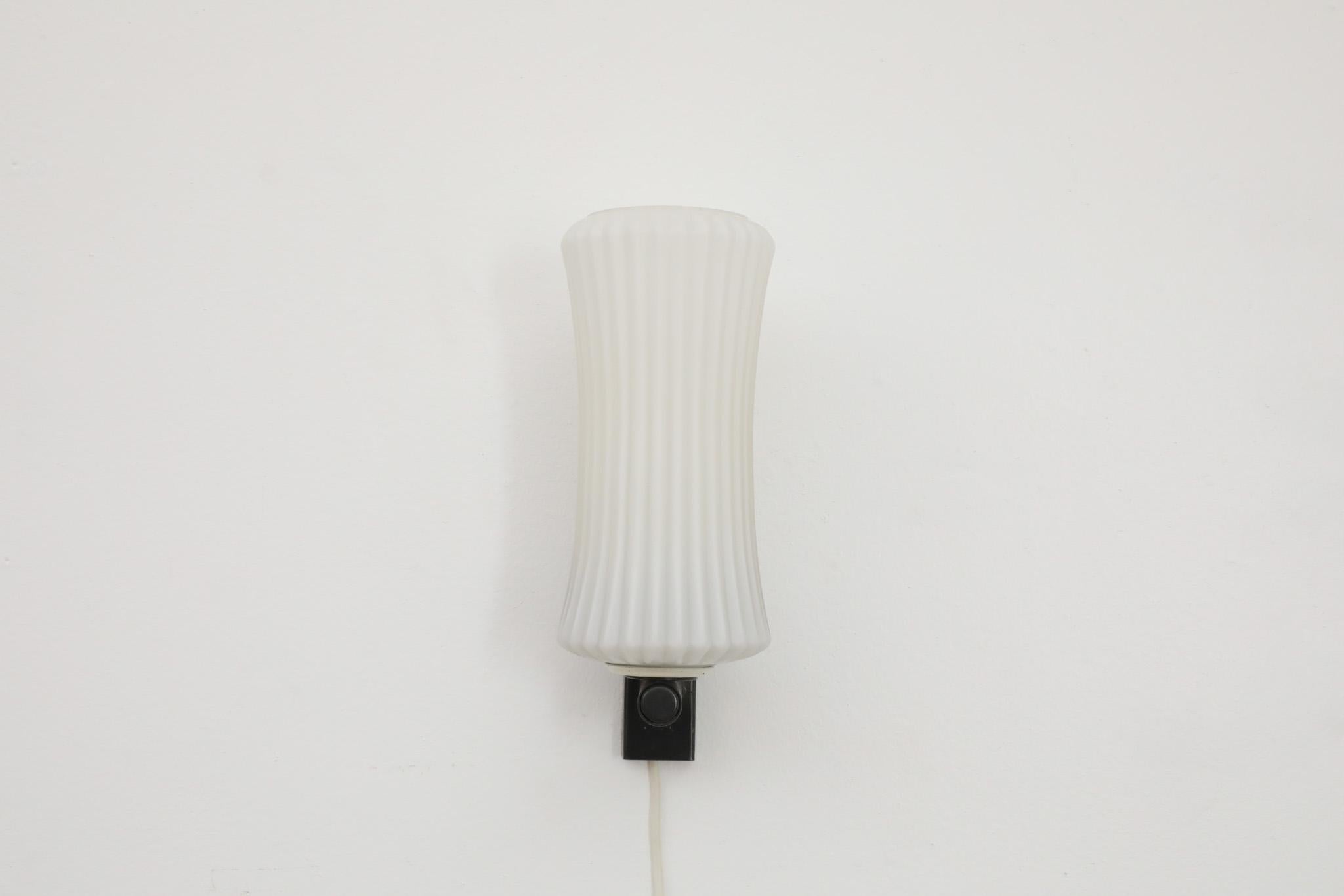 Wall mounted, Mid-Century milk glass sconce by pioneering Dutch manufacturer Philips. Has a black enameled metal base with corseted milk glass shade. The light is in original condition with some visible wear consistent with age and use. Similar pair