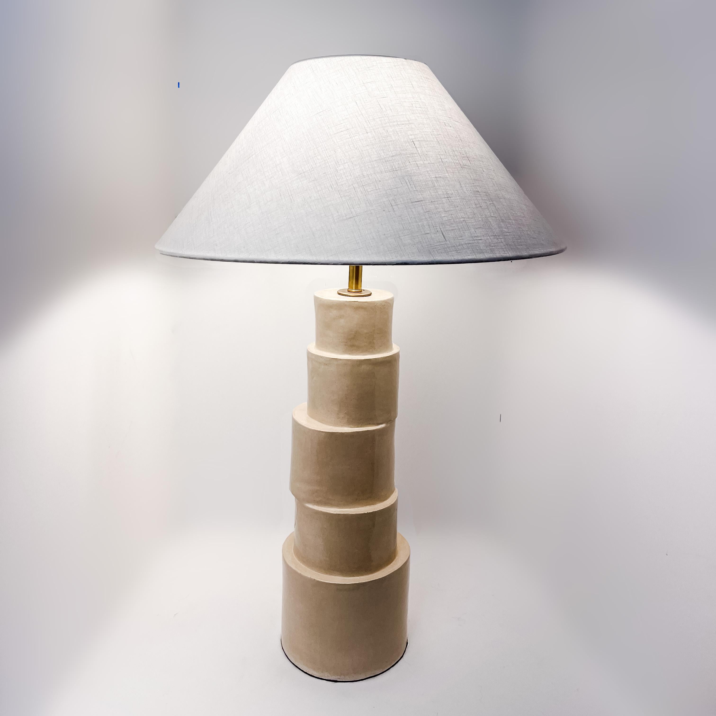 La lampe de table Stacked Column est fabriquée à la main en argile raku dans une glaçure camel avec des composants en laiton, un cordon en tissu argenté tressé et un abat-jour en lin ivoire. La nature répétitive, le ton neutre et le design