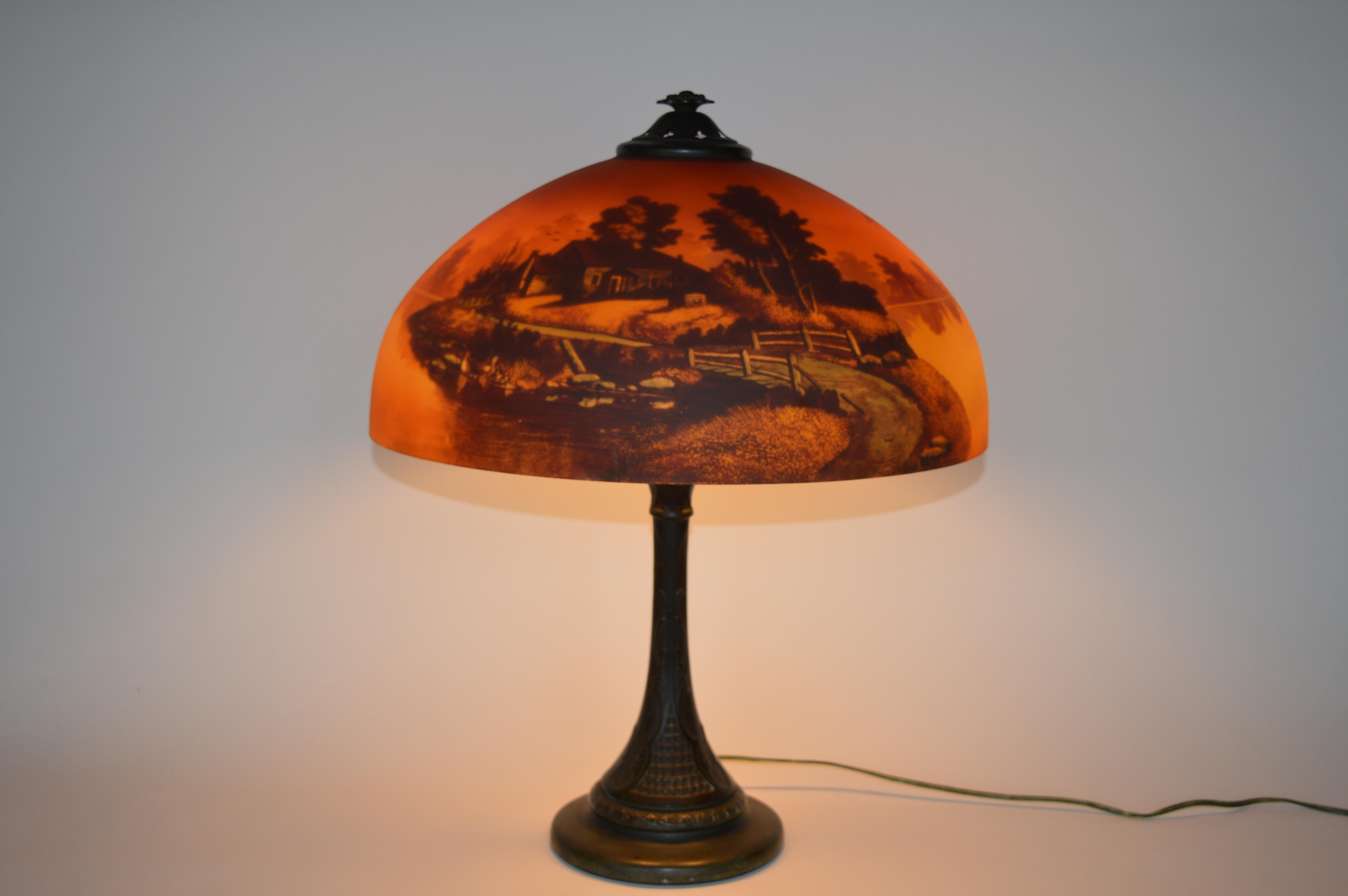Lampe de table paysage coucher de soleil Phoenix,
vers les années 1920, abat-jour en verre peint à l'envers avec une base en métal patiné. L'abat-jour représente un paysage rural de chalet au coucher du soleil. 21 pouces de hauteur totale,