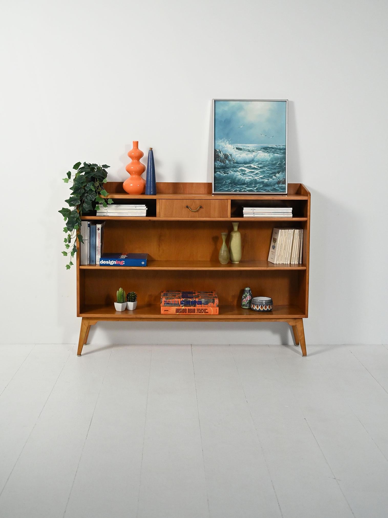 Libreria in olmo anni ’50, poco profonda e a giorno, con piedini affusolati e cassetto centrale.

Linea nordica anni ’50 in olmo dalle tonalità miele, con struttura a giorno e profondità contenuta che accentua la leggerezza visiva. Piedini svasati