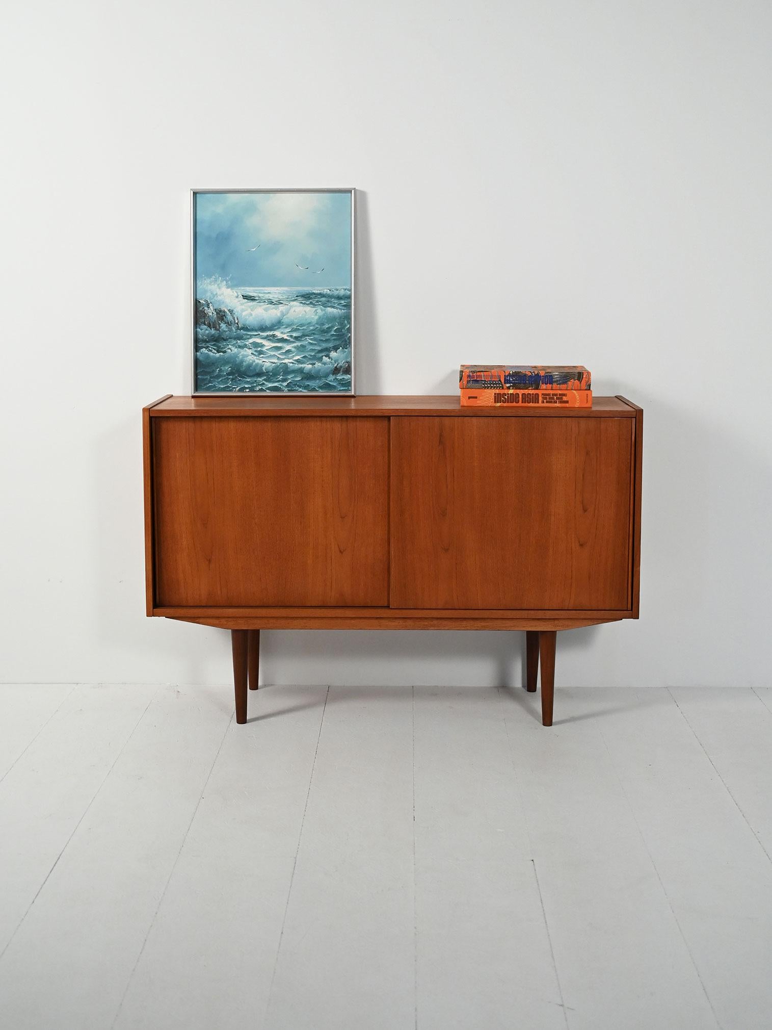 Sideboard compatto in teak con gambe affusolate, ante scorrevoli con maniglie integrate a rilievo e vano con doppia mensola.

Sideboard scandinavo anni ’50, cassa in teak con venatura calda e gambe coniche in rovere massello che alleggeriscono il