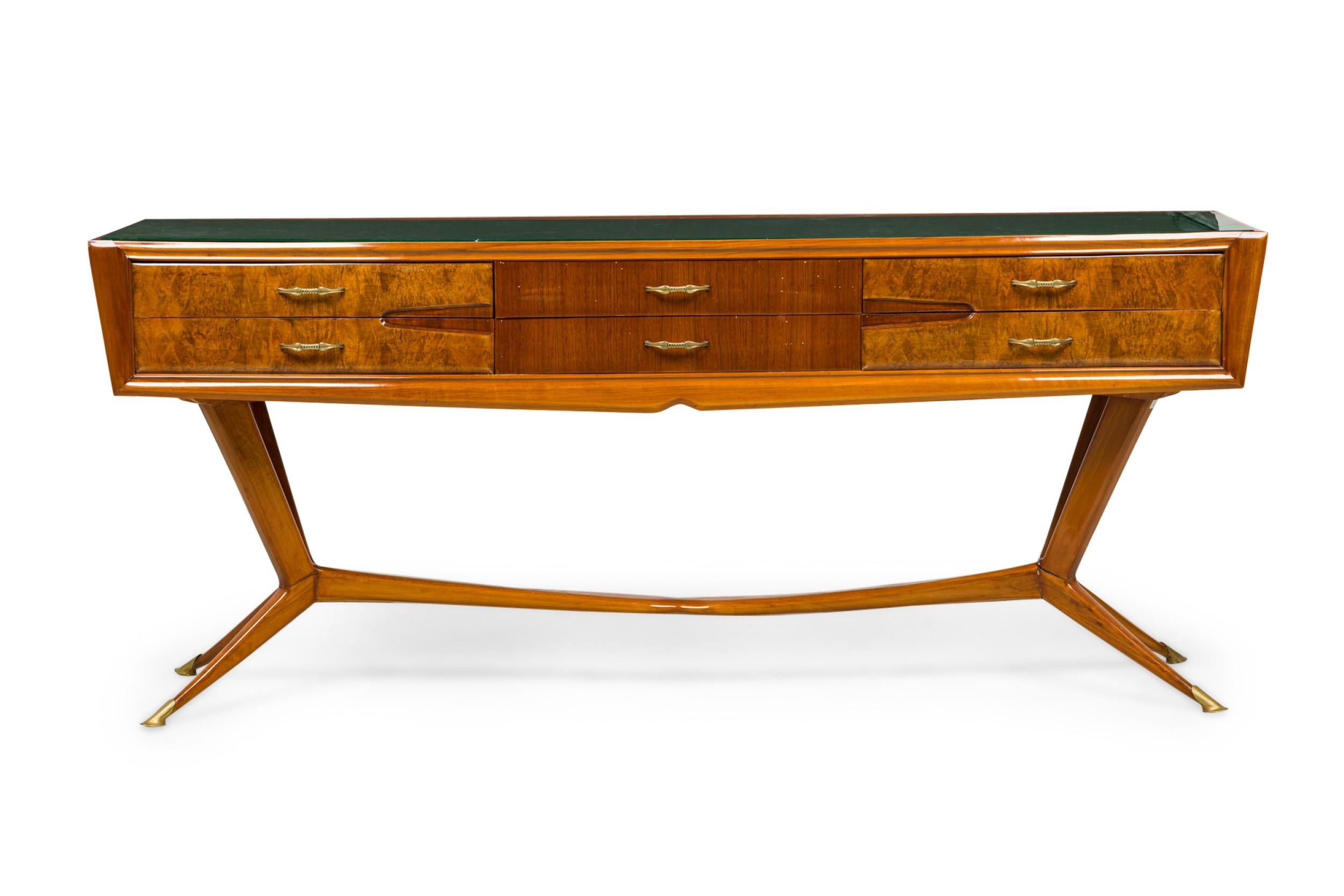 Italian Mid-Century (1954) buffet / console table with a wood cabinet featuring contrasting decorative wood veneers, 6 drawers with brass drawer pulls, topped with a green glass top with gilt detail, and resting on four angular legs joined with a