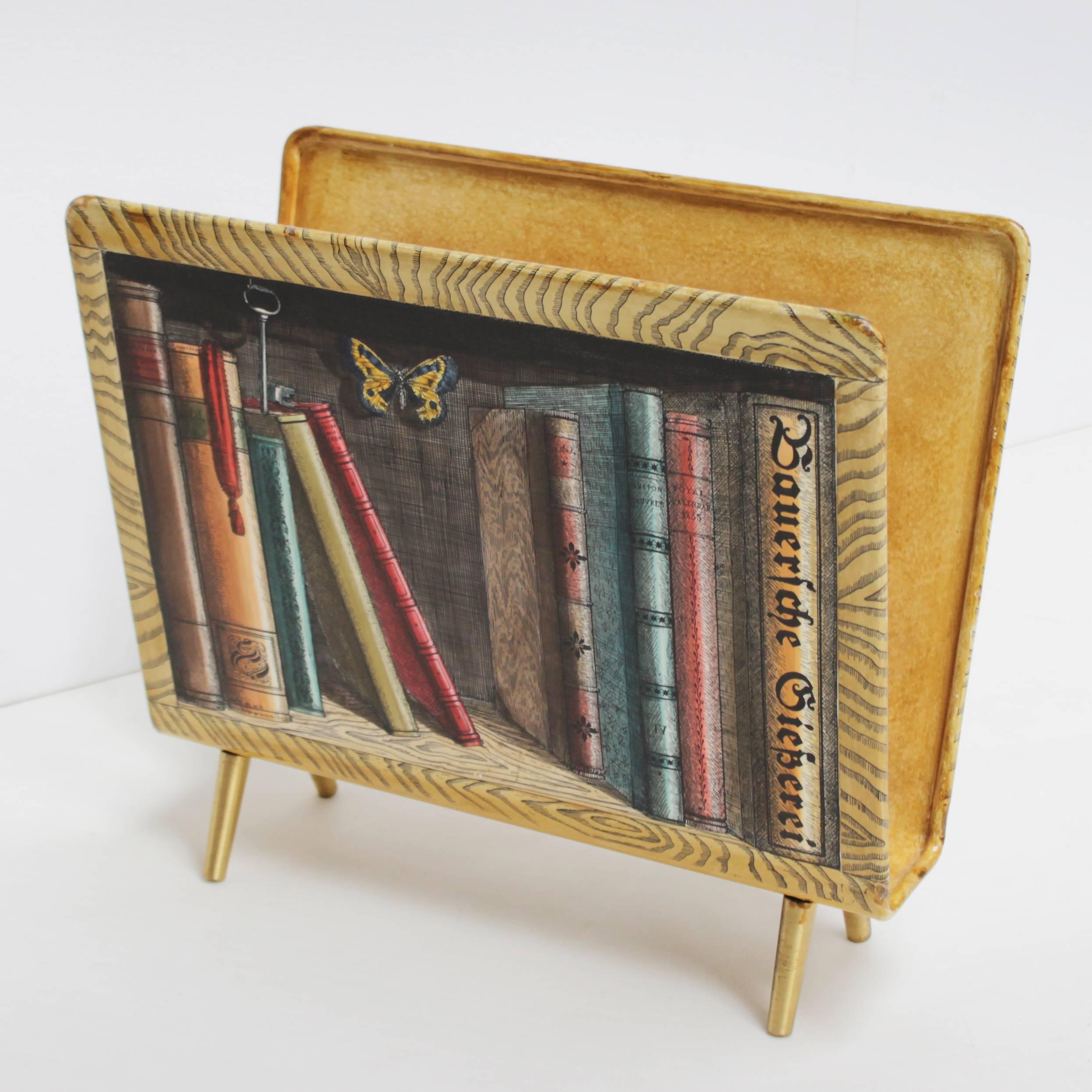 A magazine rack from the fifties by Piero Fornasetti decorated with a trompe l'oeil bookcase. This piece is lithographically printed and hand-painted in beautiful colors. 
Brass divider and feet. It's really in a top condition: no crack, no