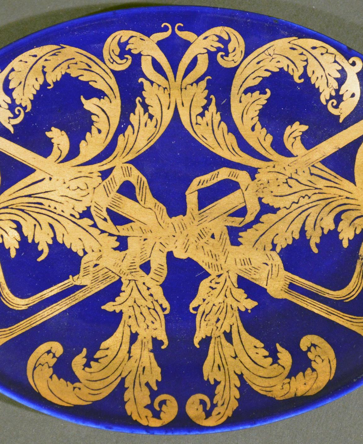 Die ovale Schale mit Pip-Motiv von Piero Fornasetti ist auf dunkelblauem Grund mit einem vergoldeten Muster aus zwei Pfeifen bemalt, die von Tabakblättern umrankt und mit einem Band in der Mitte zusammengebunden sind.

Referenz: Fornasetti: The