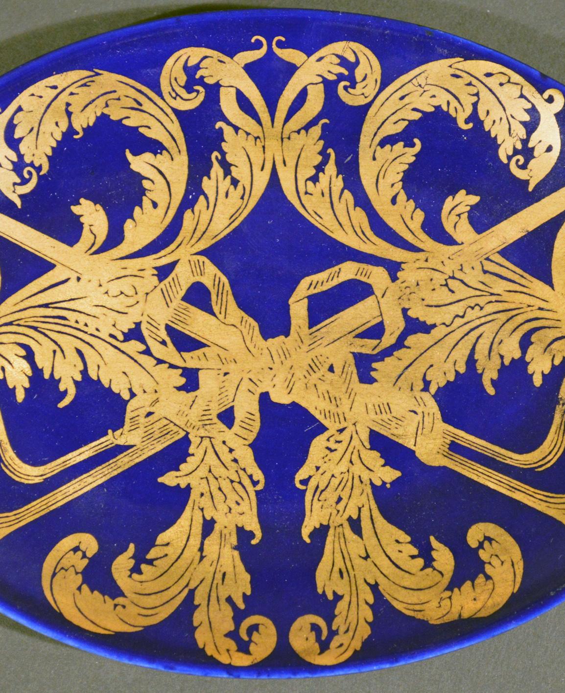 Le plat ovale à motif Pip de Piero Fornasetti est peint sur un fond bleu foncé avec un motif doré de deux pipes entrelacées avec des feuilles de tabac et attachées ensemble par un ruban central.

Référence : Fornasetti : The Complete Universe,