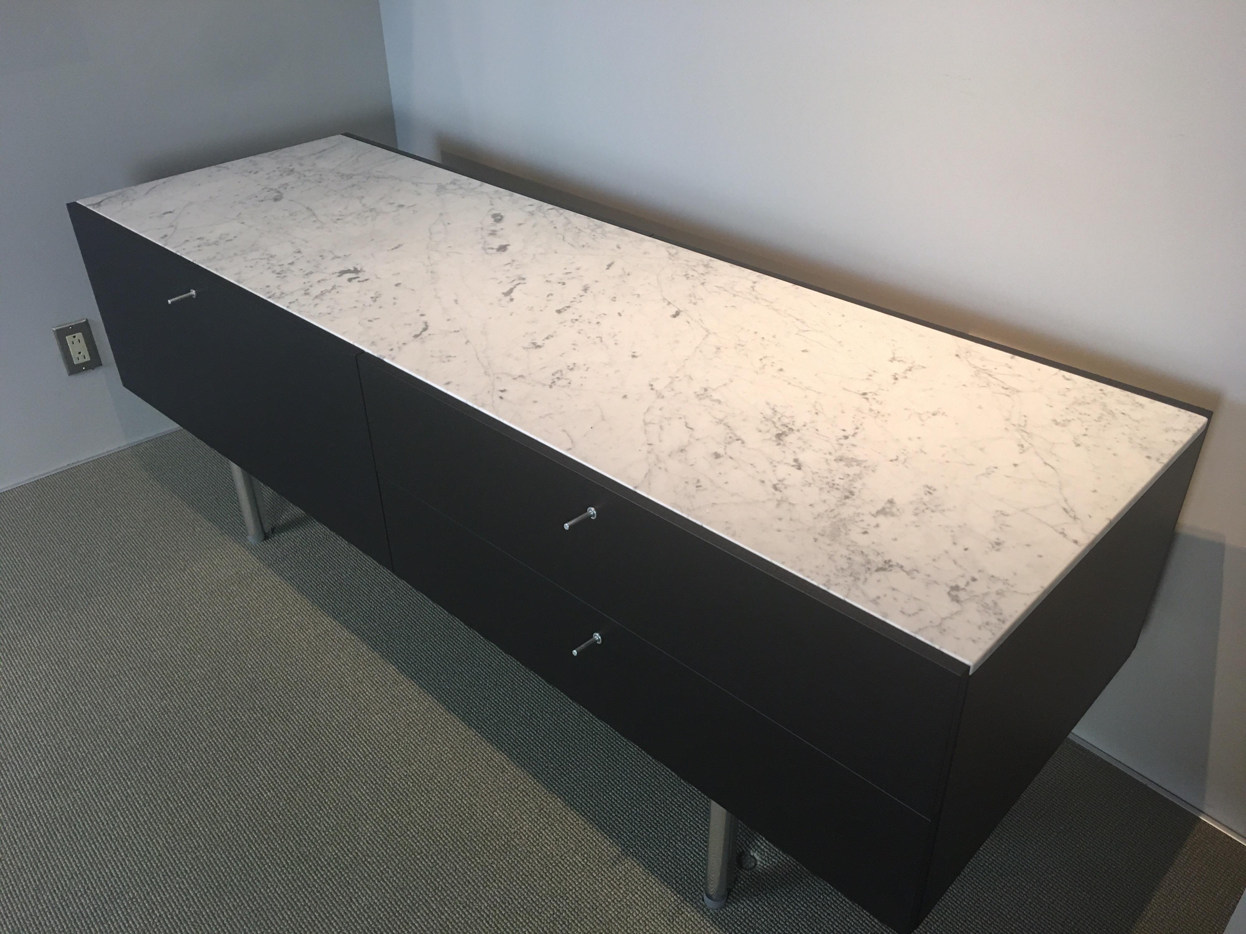 Contenitore con finitura nera tinta. Serie di piatti di Piero Lissoni per Cassina. La credenza è composta da un cassetto grande, perfetto per le bottiglie etc., e da due cassetti più piccoli. Il piano in marmo bianco si trova all'interno del labbro