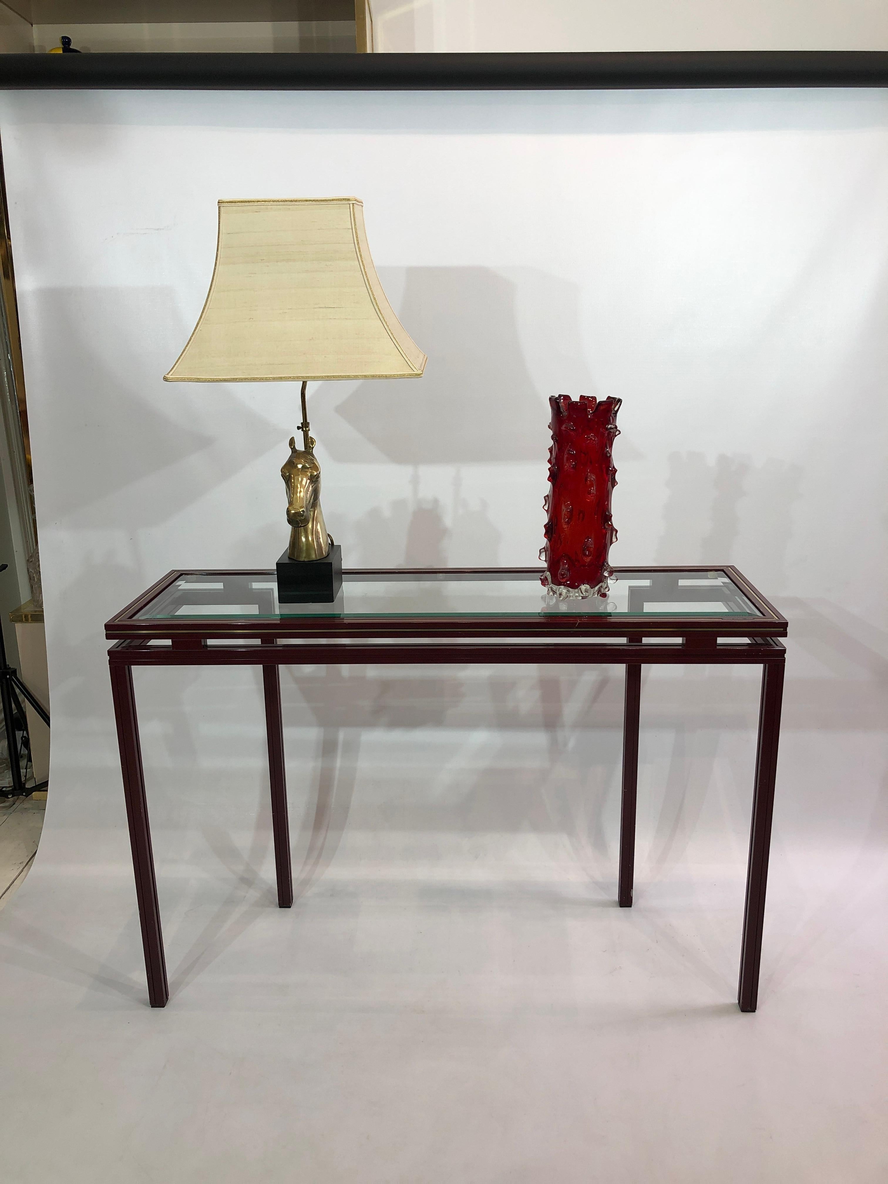 Burgundy console table with clear glass top on an interesting juxtaposition front to back effect base. The base is made of aluminium and powder coated in burgundy and finsifed with the characteristic Pierre Vandel brass inlay. In good vintage