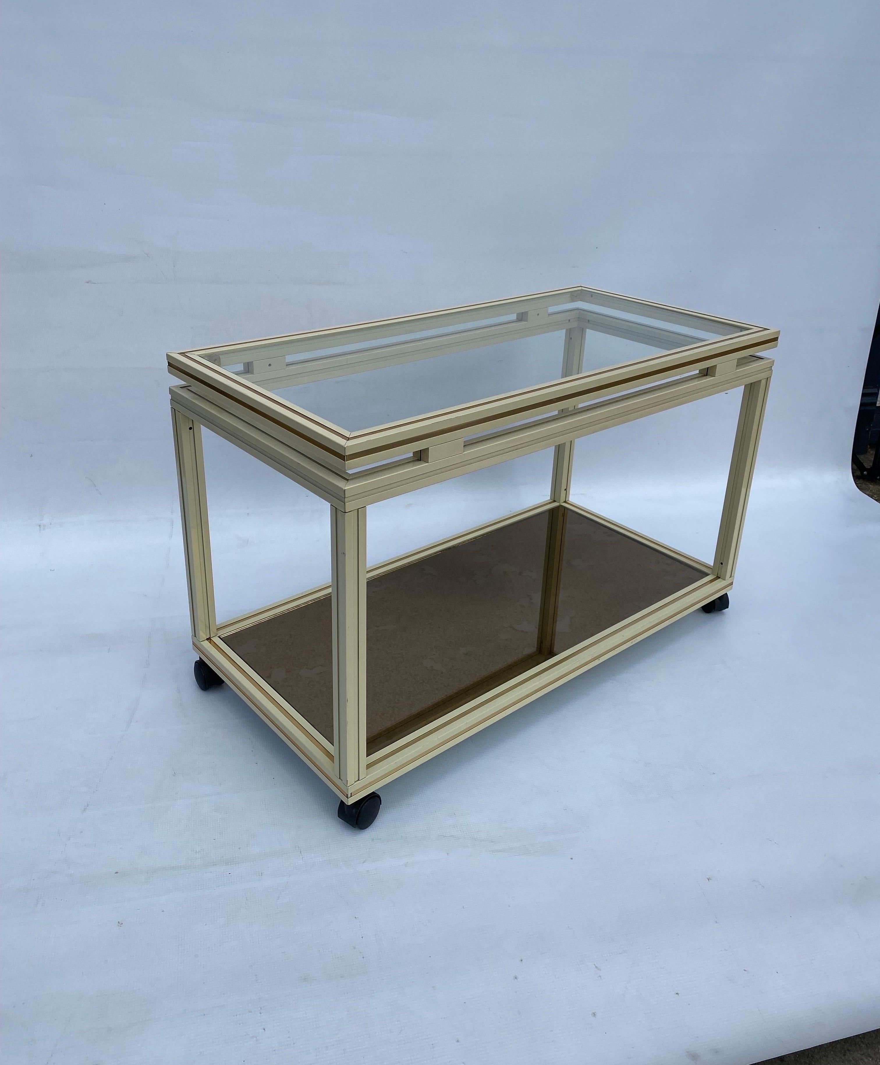 Cream and brass trimmed drinks cart or side table on wheels by Pierre Vandel. Clear glass top and a smoked antique mirror on the bottom is just a great and practical piece. This desirable piece is an easy one to navigate around and decorate it in