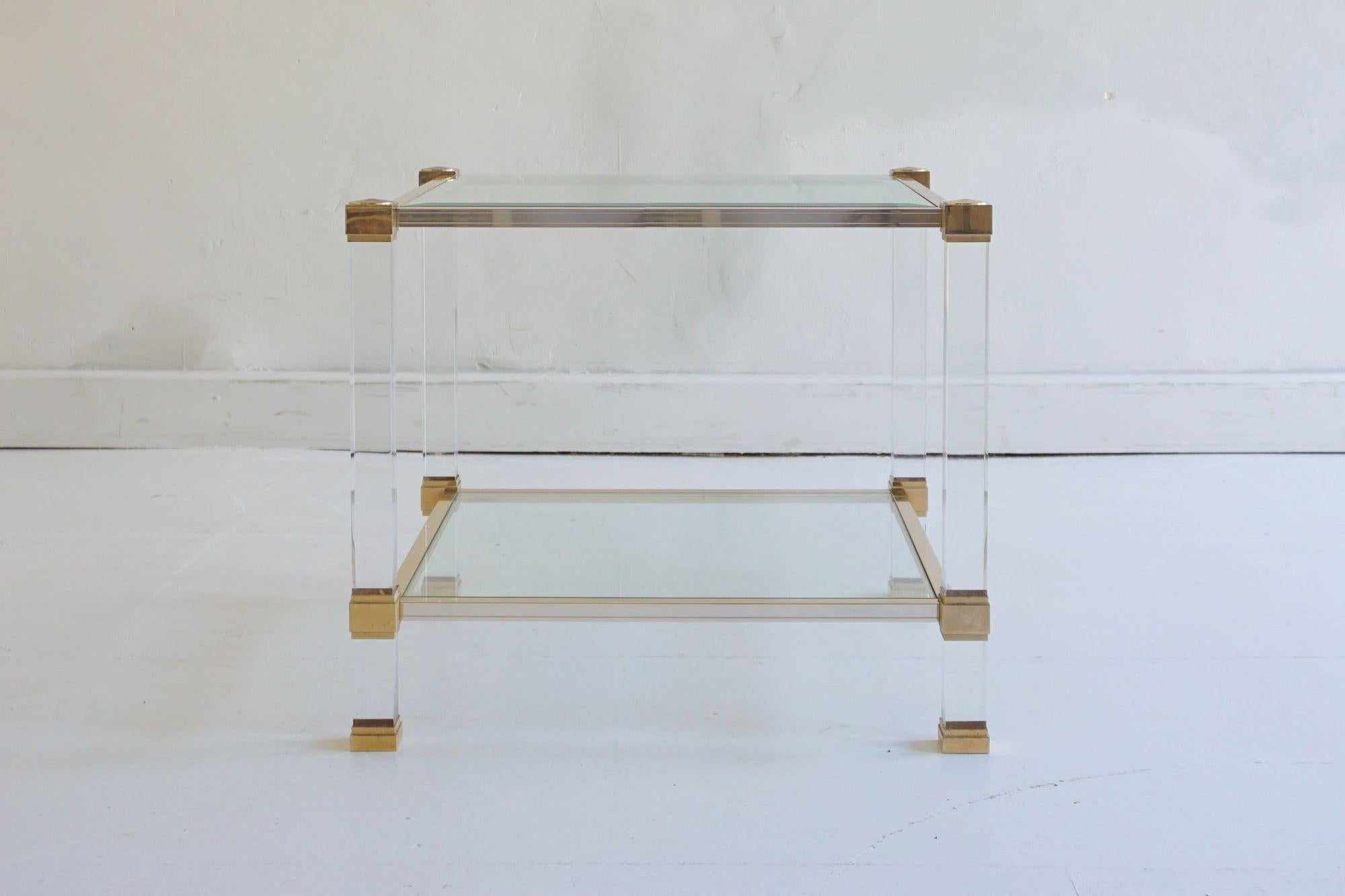 This two-tier side table by Pierre Vandel features a clear Lucite frame paired with a gold metal structure for a clean, modern look. The table includes two glass surfaces, the top glass with beveled edges that add a subtle touch of detail. With its