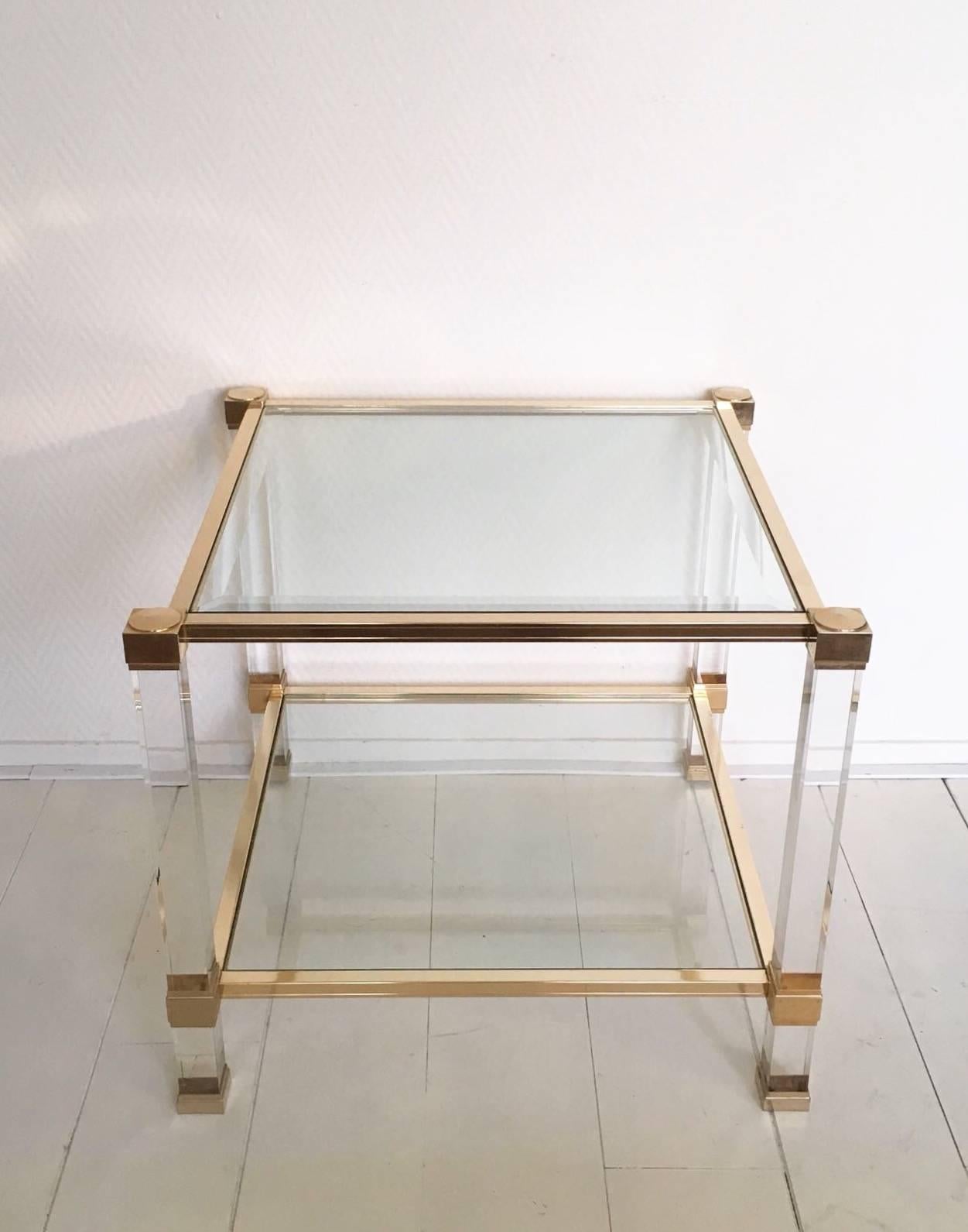 This two-tiered small coffee or side table was designed by Pierre Vandel Paris. It features a gold coloured metal base with beveled glass and partly Lucite feet. The table wear's the name of it's designer and remains in wonderful condition. Très