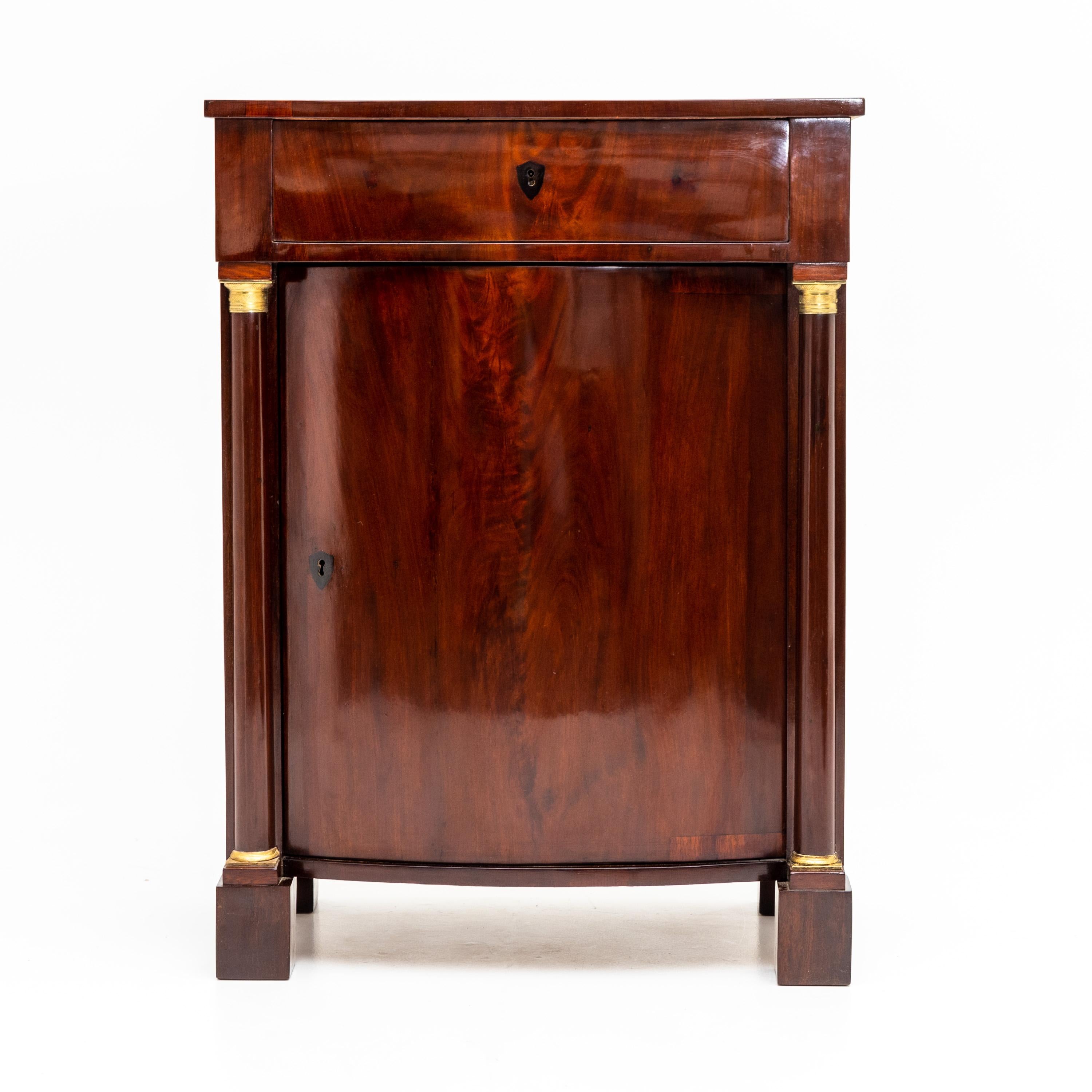 Pillar cabinet with cambered body, one drawer and door, and pillar supports with brass bases and capitals. Mahogany veneered.