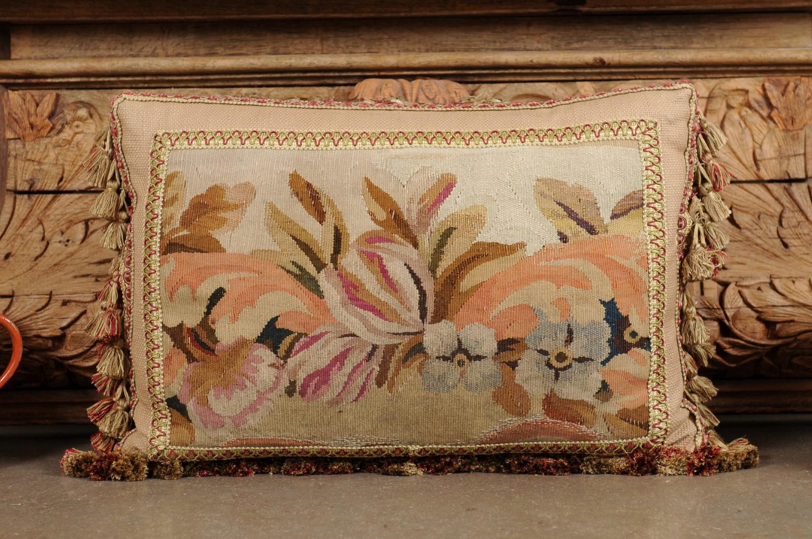 A pillow made from 19th century French tapestry, with floral decor and petite tassels. Created from a tapestry woven during the 19th century, this horizontal pillow is adorned with a delicate decor of blue and pink flowers accented with green, brown