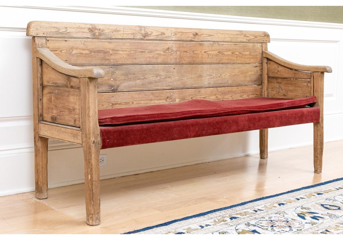 A very well made Pine Bench constructed from Antique wood with beautiful graining, planked back, paneled sides, gently sloping arms and a fine Rustic presentation. The wood has an overall soft feel and there is a Burgundy fabric seat cushion with