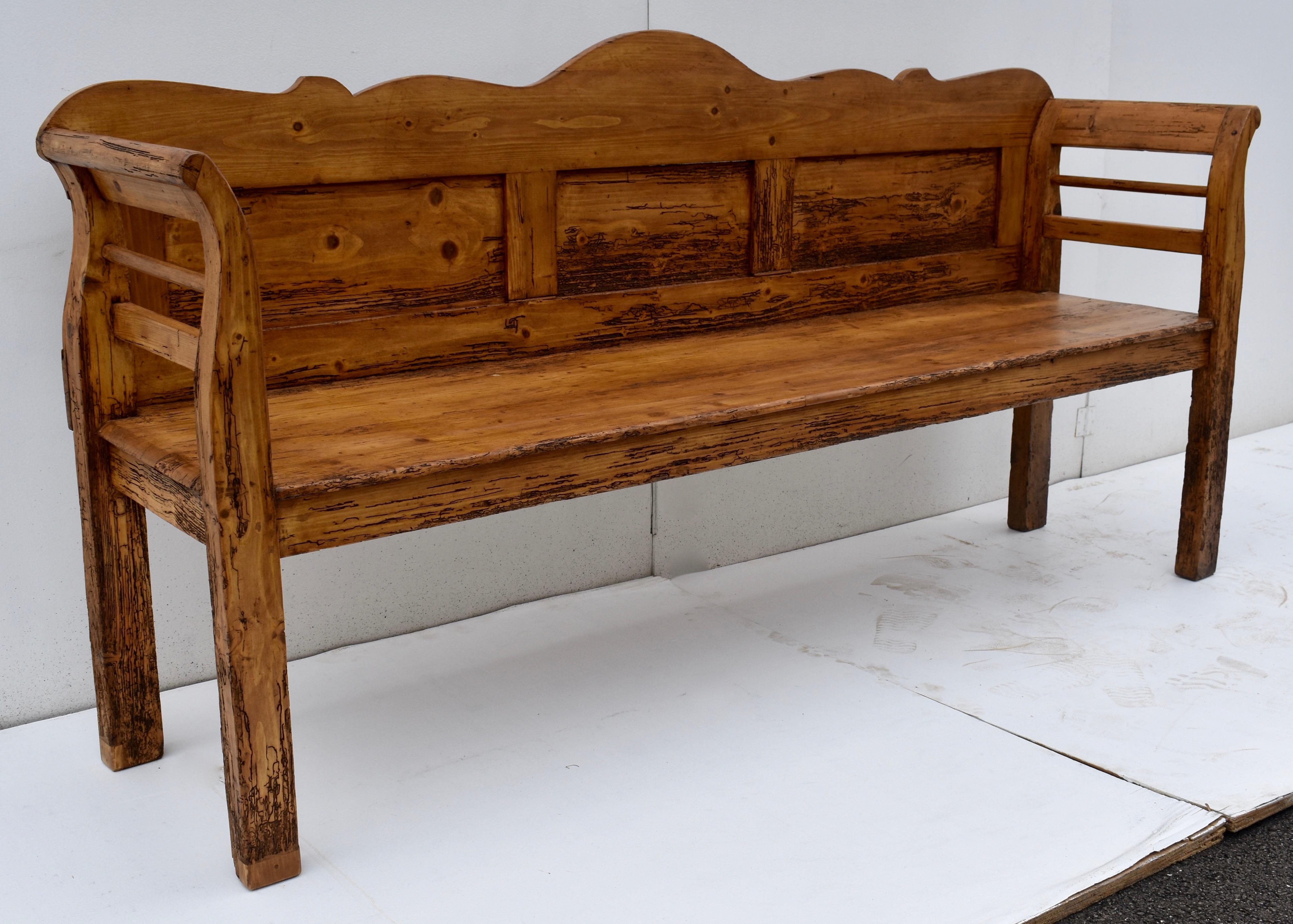 This long, low pine bench or settle has a gently scalloped camel back above three flat panels. The back is fixed to the rear of the combined arm and leg which is nicely scrolled at the top with sraight legs to the floor. The deep seat is braced all