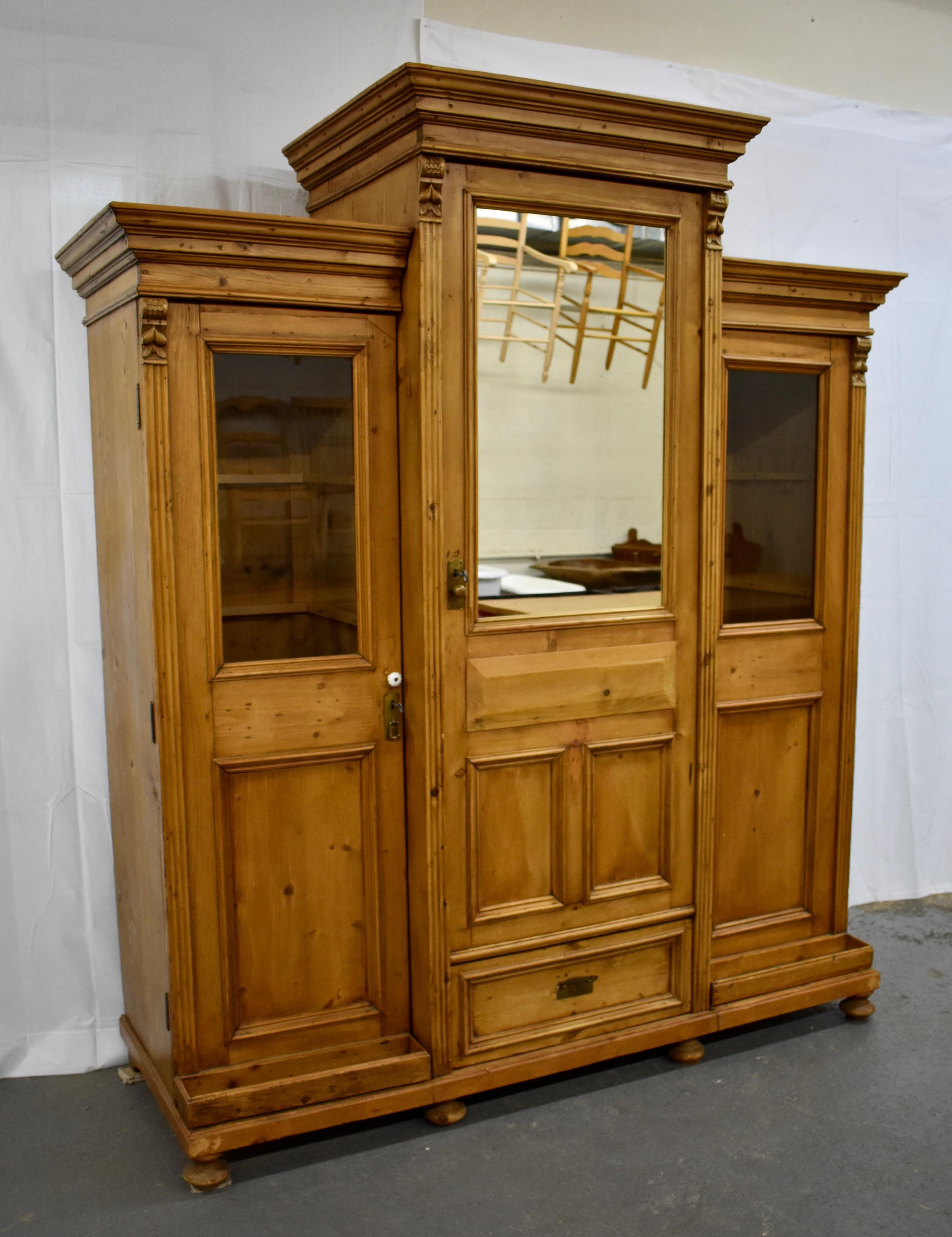 This large pine breakfront hall wardrobe features a tall central section with a bold crown molding, a half-mirrored door with a false drawer front and two flat panels, and a deep handcut dovetailed drawer. This is flanked by side sections, the doors