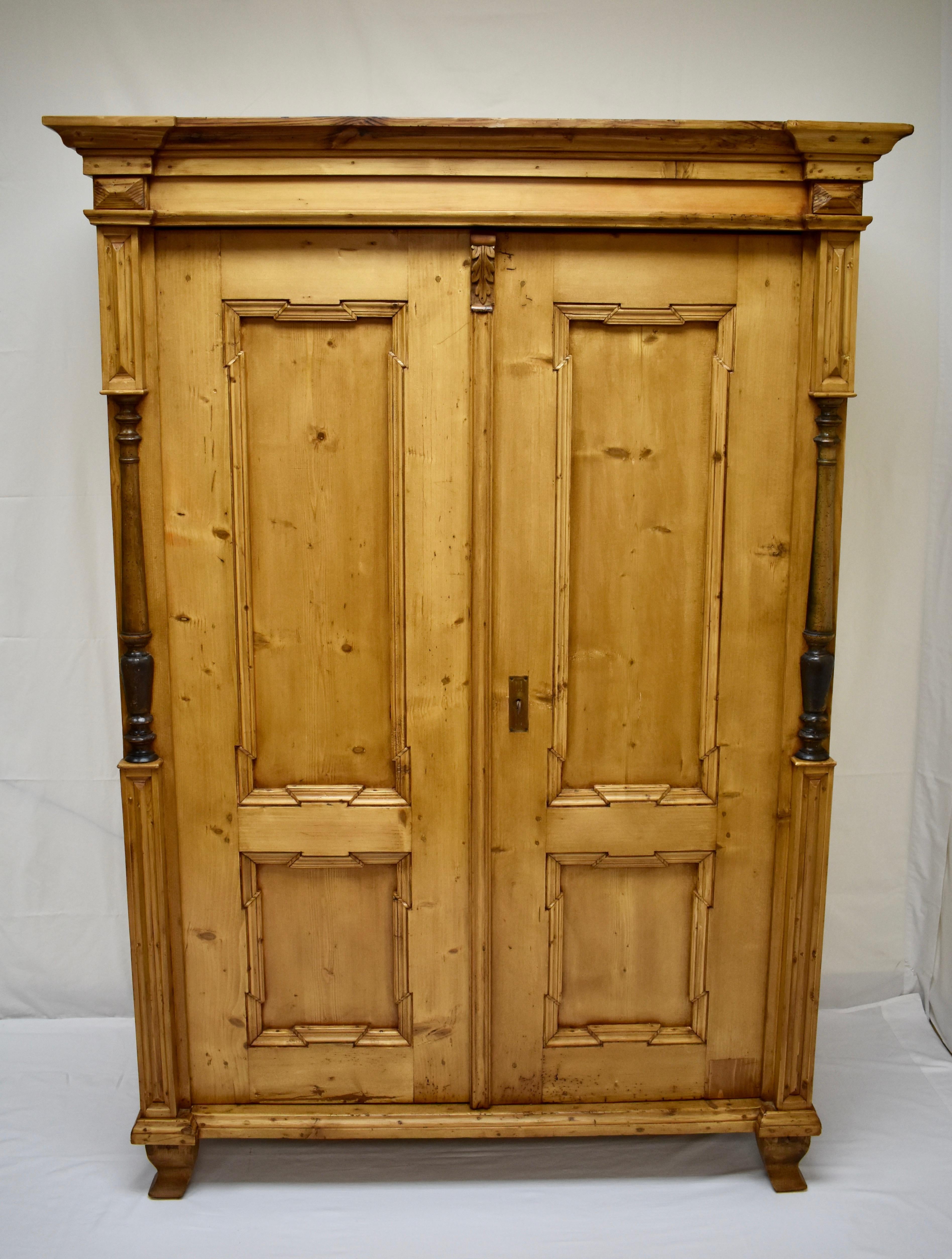 This handsome pine armoire features a bold crown molding above two doors, each with two geometric panels, separated by a fluted strip capped with a hand-carved acanthus leaf corbel. The front corners are decorated with ebonised beechwood split