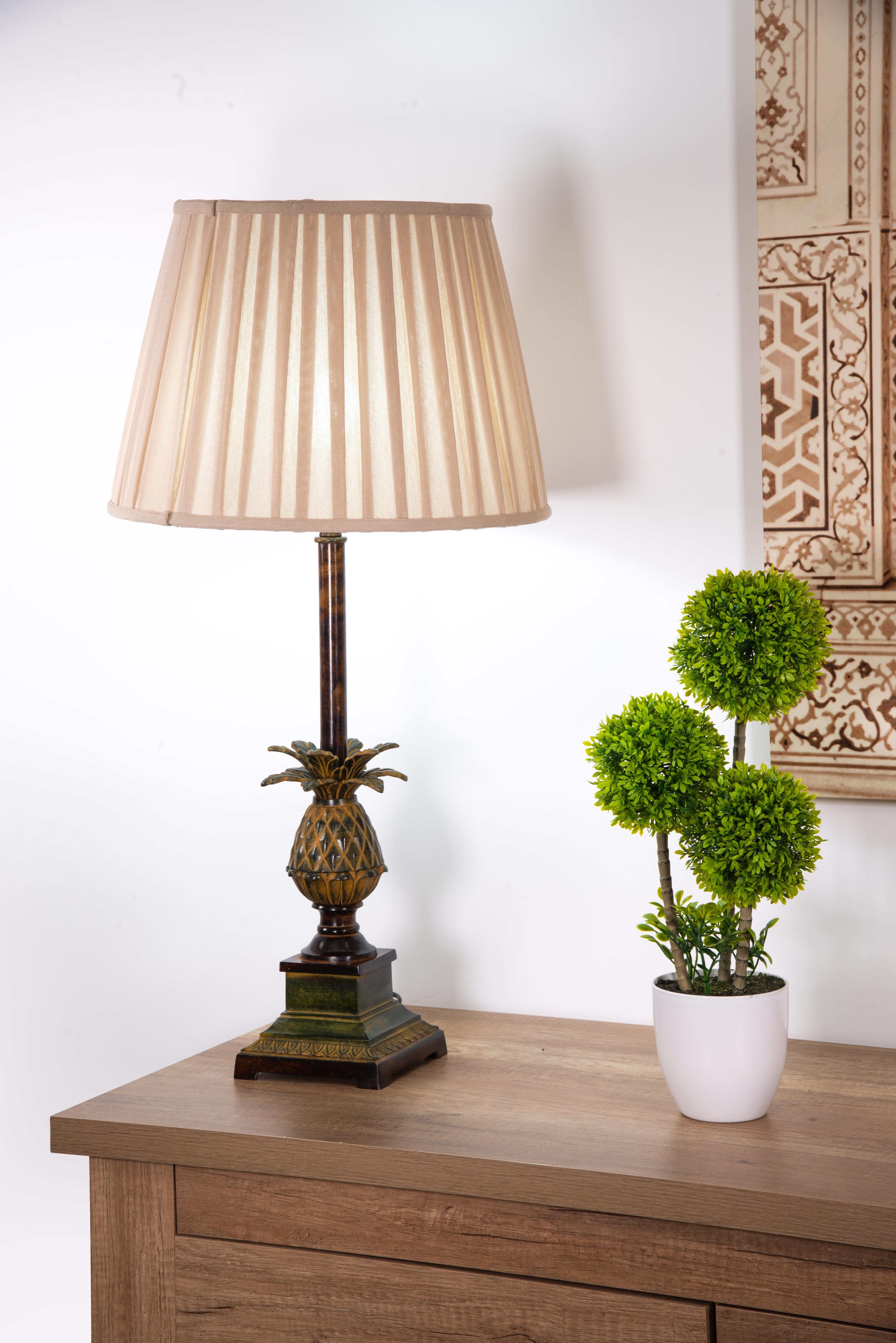 Lámpara de bronce patinado. 
Sin pantalla. 
Una base cuadrada bajo un pedestal resalta el curioso elemento decorativo que protagoniza esta obra: la piña está formada por un cuerpo ovalado con decoración de rombos rematado por un penacho de hojas que
