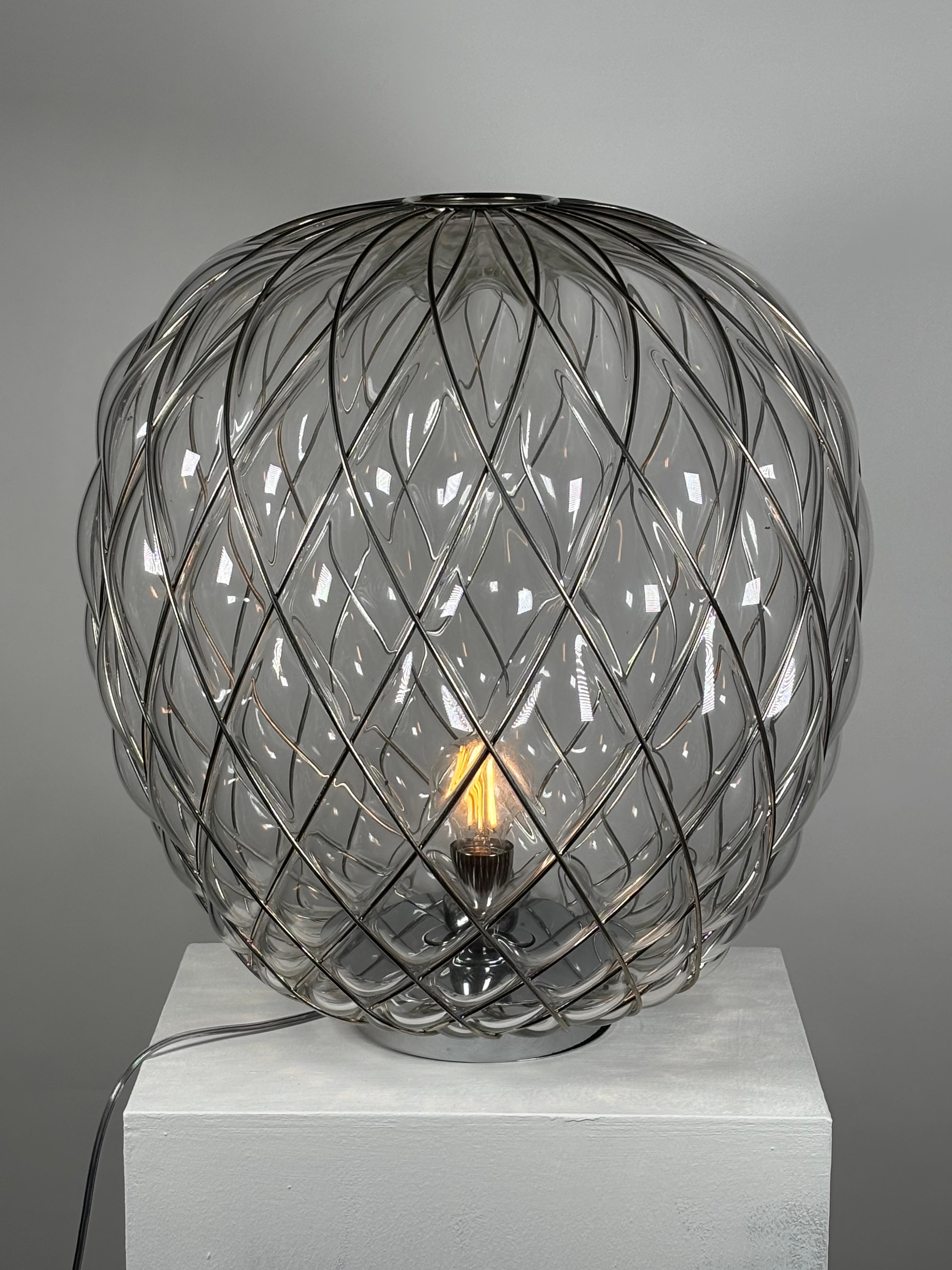Pinecone is a support lamp, characterized by a diffuser produced using the ancient technique of caged blown glass: the master glassmaker blows the glass bubble into a metal cage, which contains its natural expansion only in part. In this way the