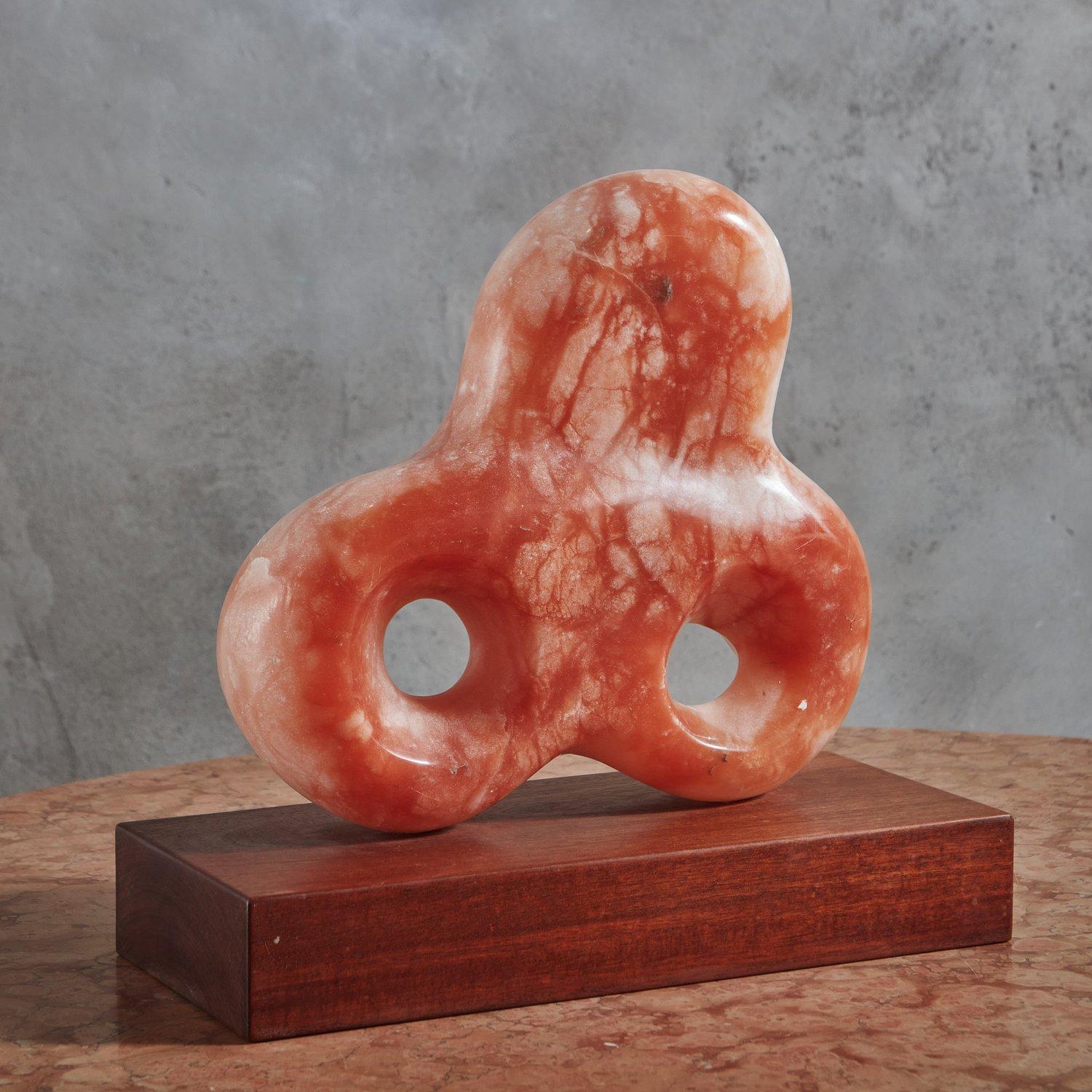 A vintage sculpture carved from a gorgeous pink alabaster stone with a range of coral, pink and salmon hues. This abstract piece features elegant curves and circular cutout details. It is presented on a rectangular wood base. Unsigned. 20th
