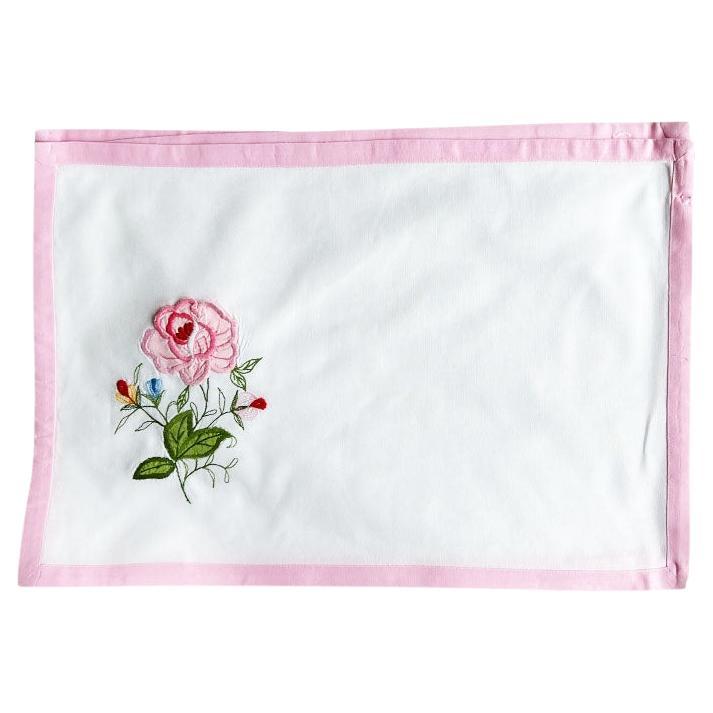 A set of four flower motif fabric placemats. Hand sewn, this set features 4 rectangular placemats in a crisp white, with a ballet pink border. Each piece is decorated with an embroidered applique of a pink English rose, with lush green leaves and