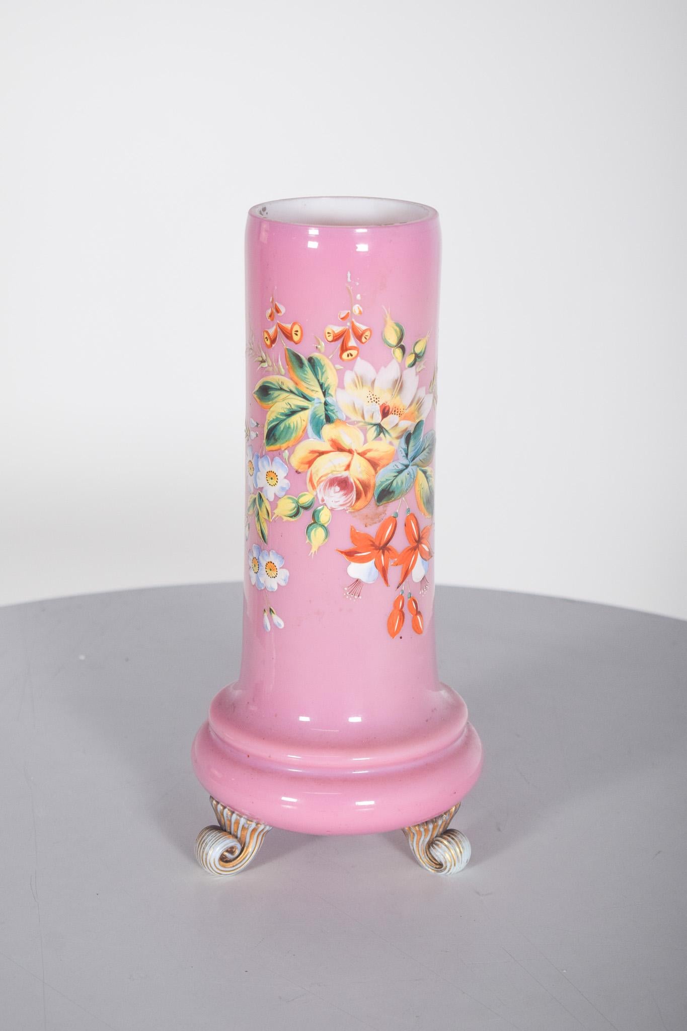 Rosa Glasgefäß. Polychrome Dekoration mit bunten Blumen, die weiße, gelbe und orangefarbene Blütenblätter hervorheben, kombiniert mit Blättern in leuchtenden Grüntönen. Kleine Details in Orange und Weiß verleihen der Komposition einen Hauch von