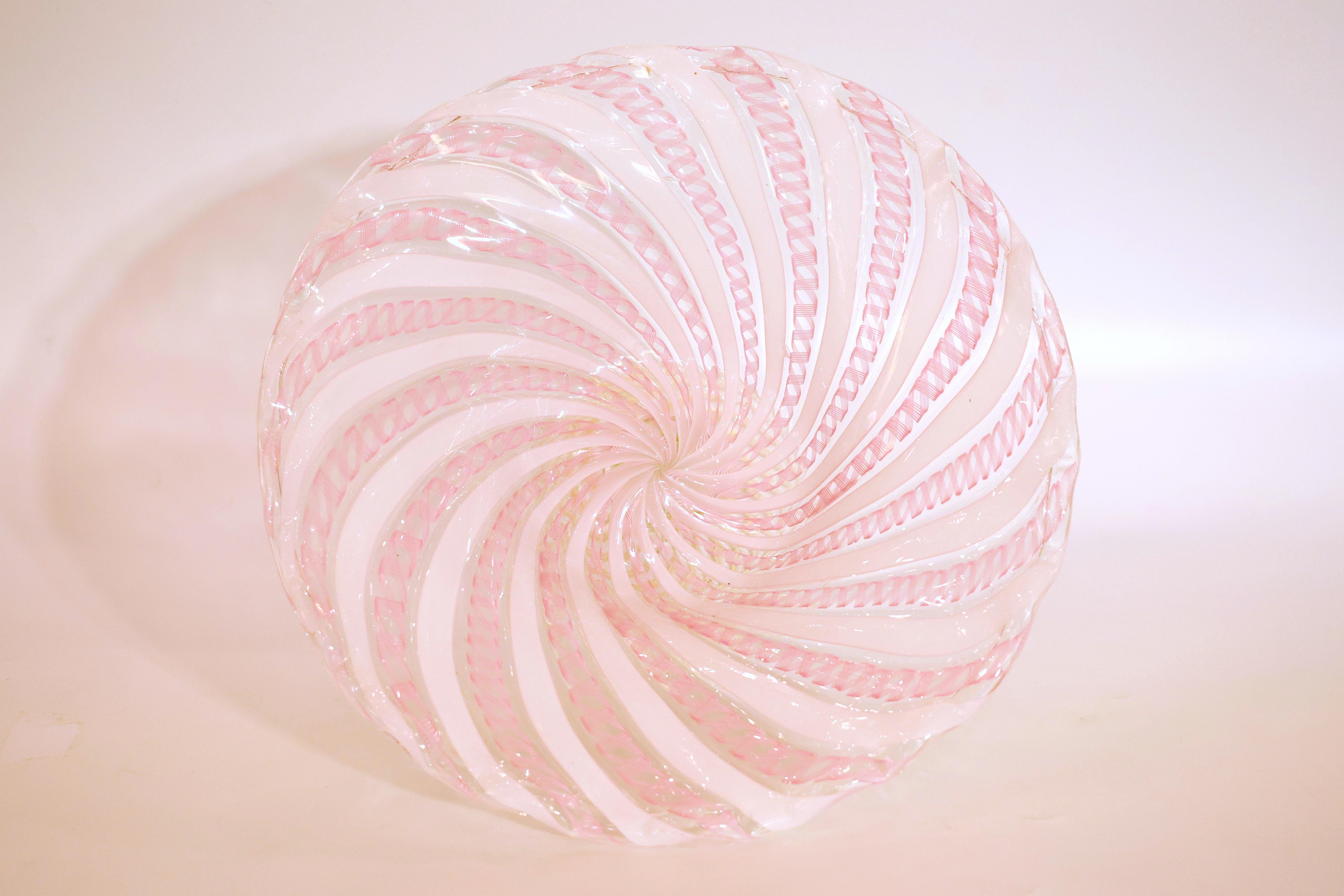 Questo centrotavola o caramella in vetro di Murano presenta la tecnica del latticino, caratterizzata da delicati motivi a nastro bianchi e rosa incastonati nel vetro. La ciotola, ampia e poco profonda, è sostenuta da un piedino a spirale con