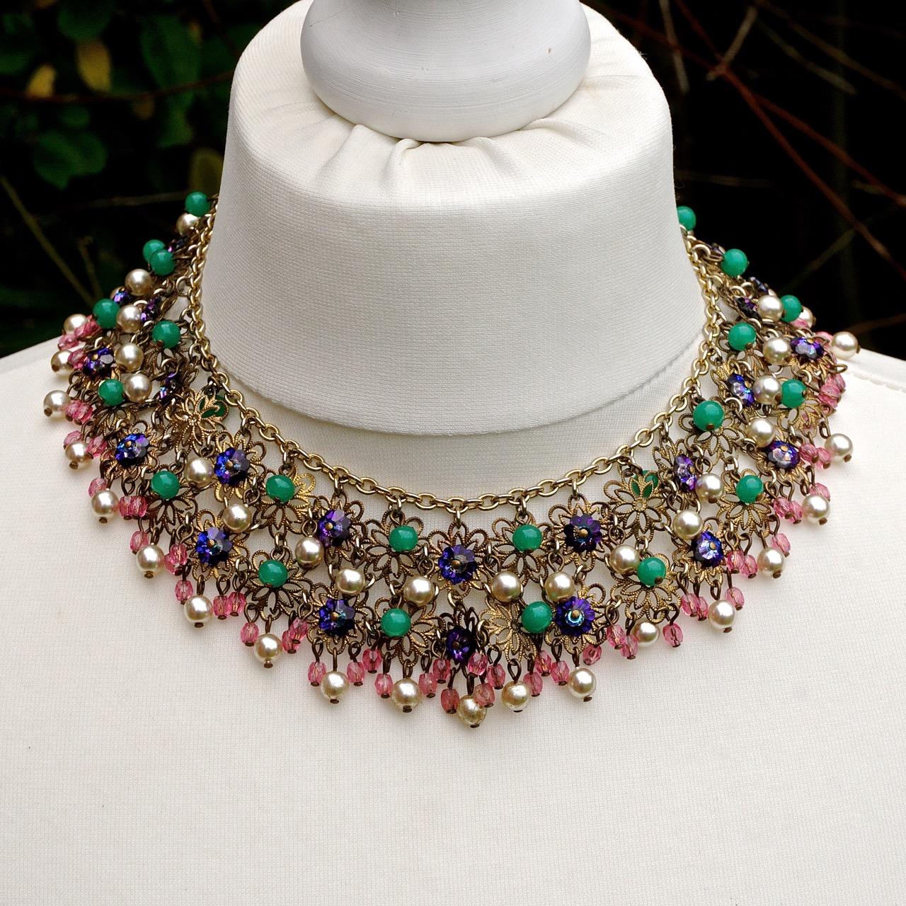 Fabulous detailed glass bead and faux pearl collar necklace. The glass beads are faceted pink beads, round green beads, purple flat beads, and faux pearl beads. They are attached to two rows of gold tone filigree flowers, which are held together at