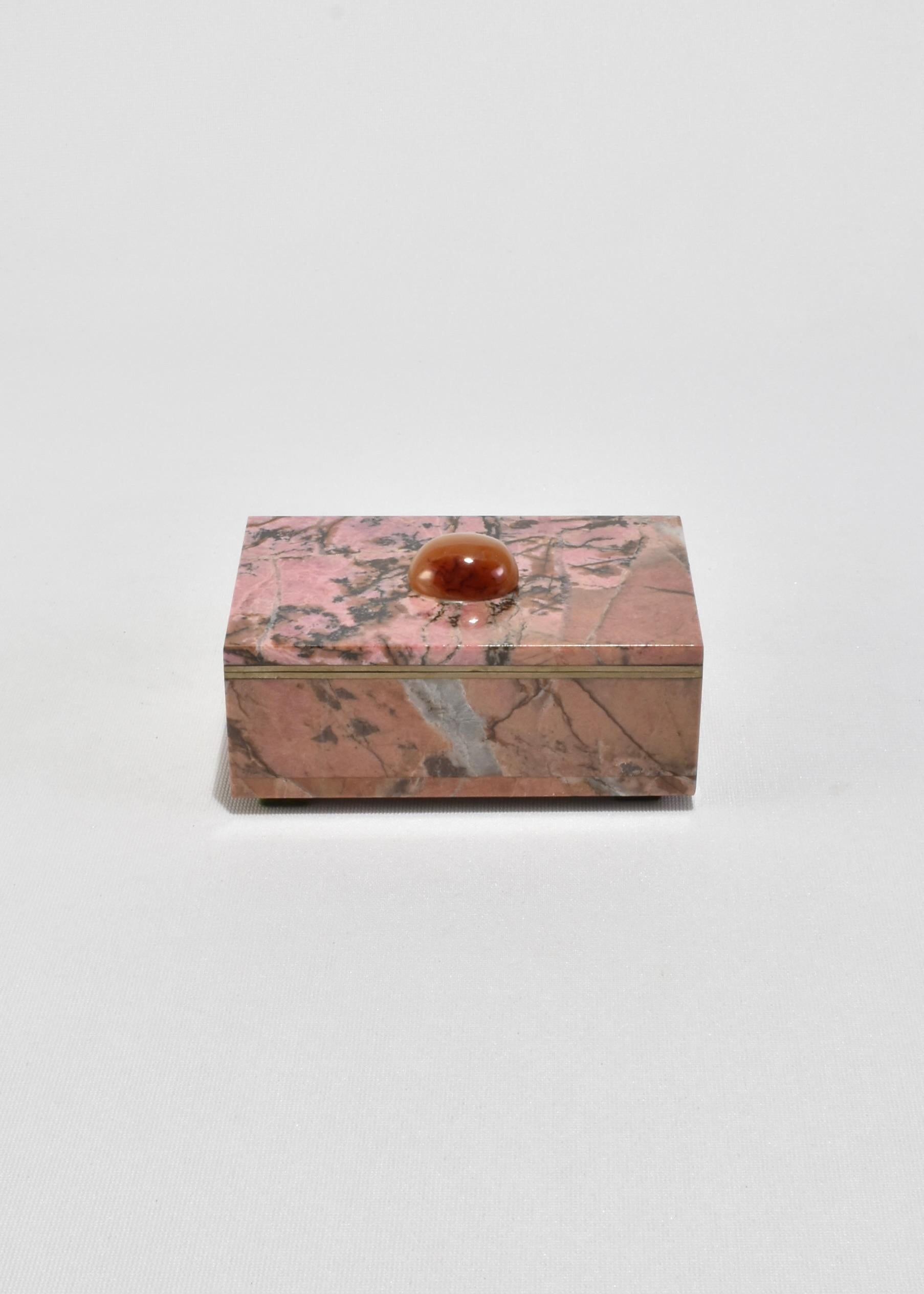 Caja vintage de rodocrosita pulida con llamativo veteado jaspeado en rosa, negro y gris, rematada con un cabujón oval liso de ágata. El interior está forrado de fieltro naranja vibrante, con adornos de latón que enmarcan la tapa y la bisagra. Las