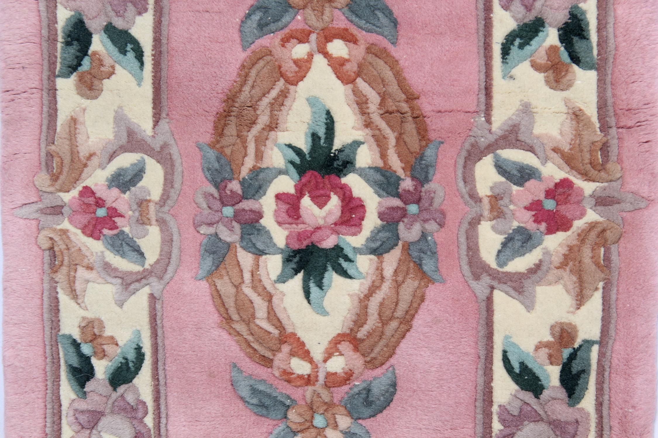 This elegant handmade carpet was constructed in the 1960s in China. The central design has been woven on a pink field with peach, purple and green accent colours that make up the floral medallion and the symmetrical surrounding design. This is then