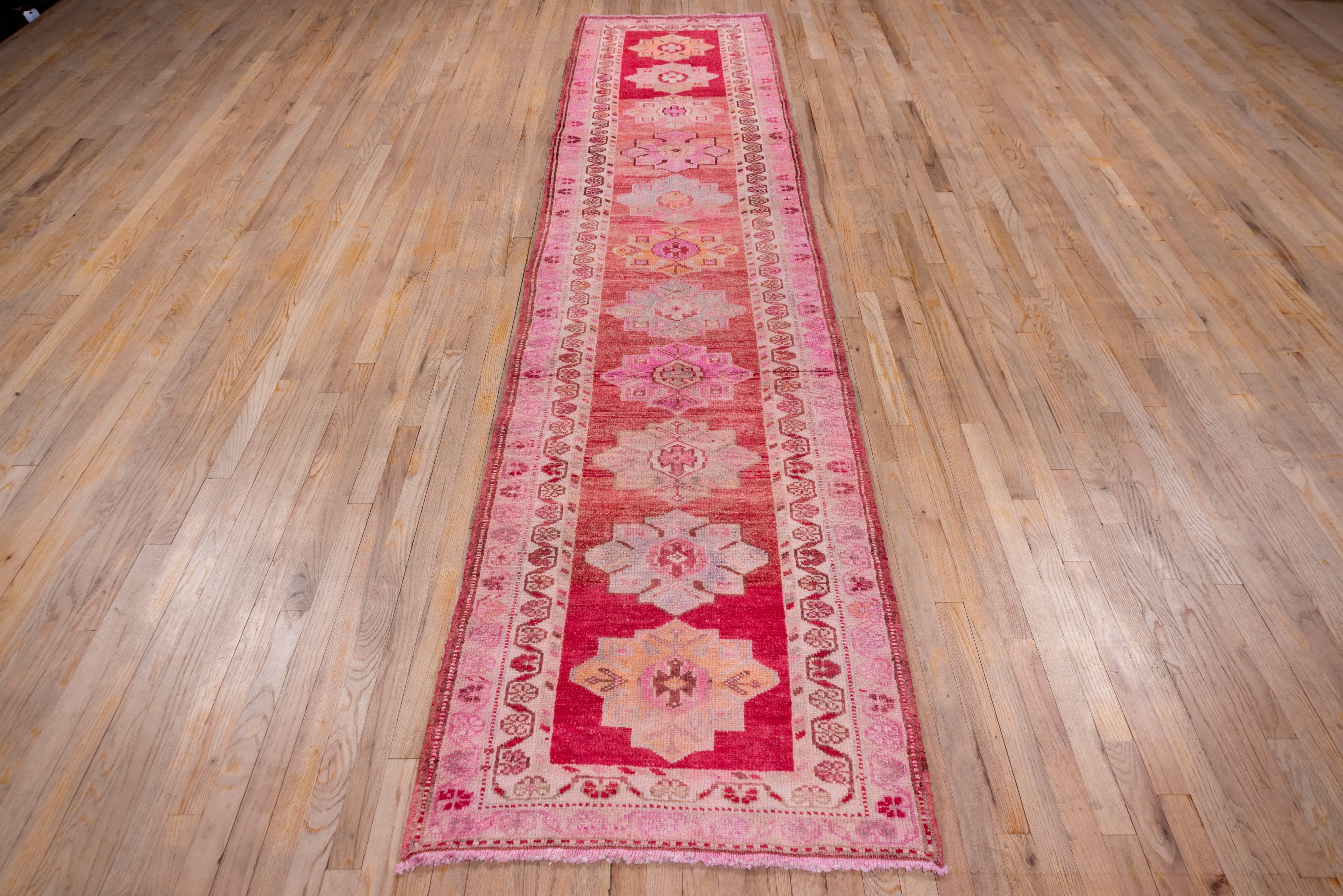 The variegated soft red field of this Anatolian displays 11 stepped diamond medallions in soft yellow, blue grey and yellow grey. The two borders both employ an undulating vine and simple palmette pattern on ivory and pale pink grounds.