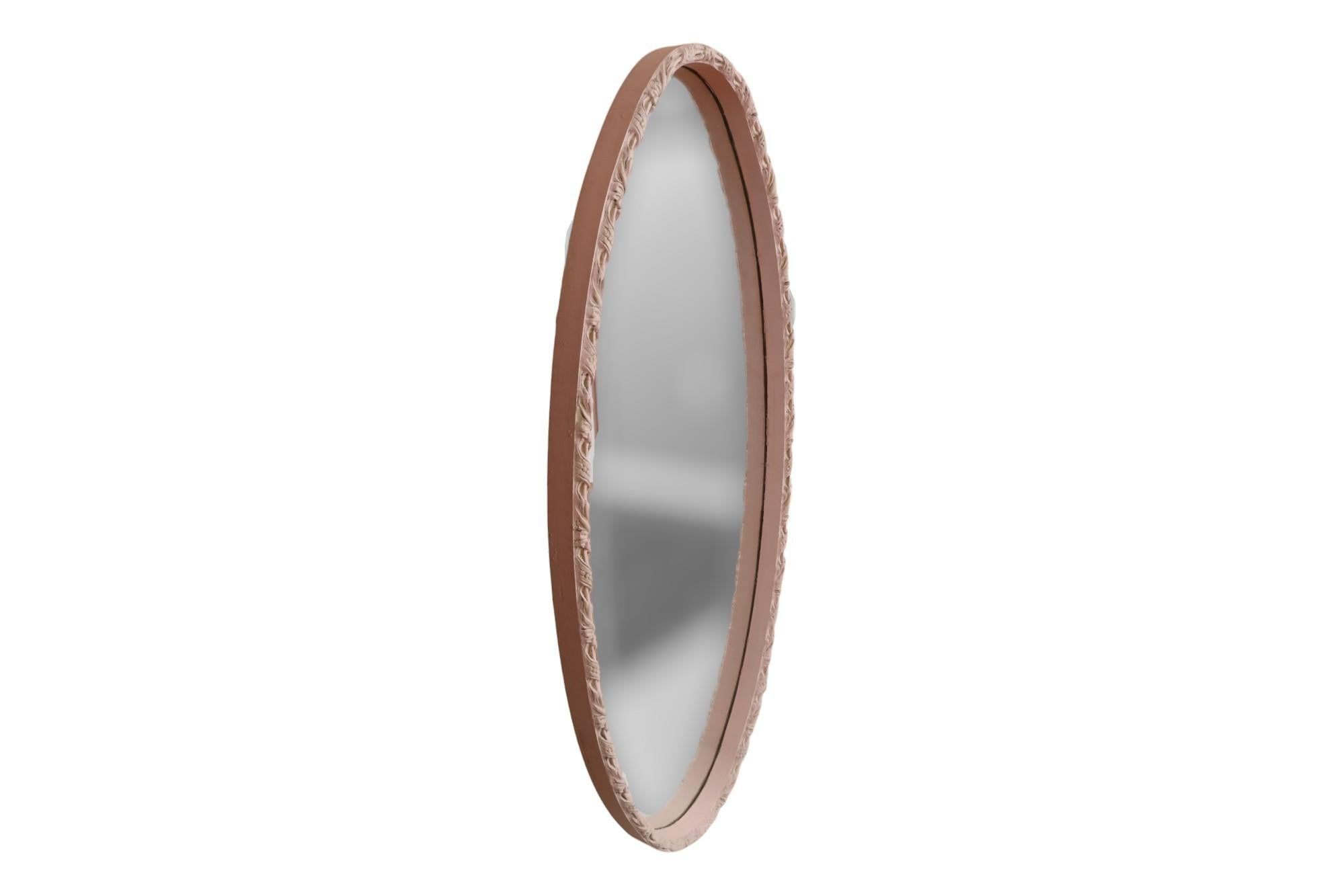 An oval wall mirror with a pink wooden frame. The deep frame is carved with a flowing ribbon motif and finished in a blush pink.