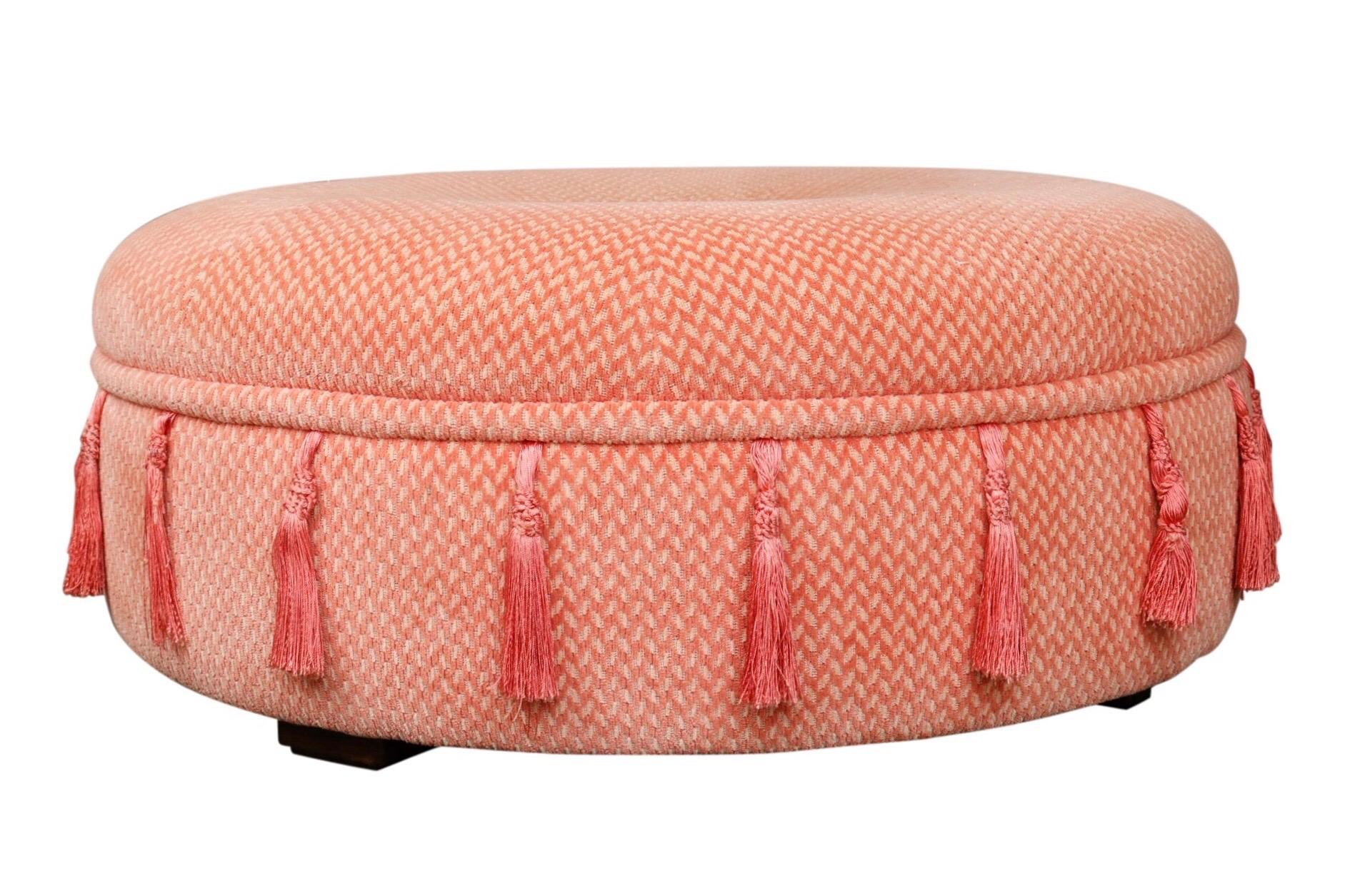 A large round coffee table ottoman. Upholstered in a salmon pink basket weave with a button at the center and trimmed with tassels around the circumference. Stands on four square wooden legs.