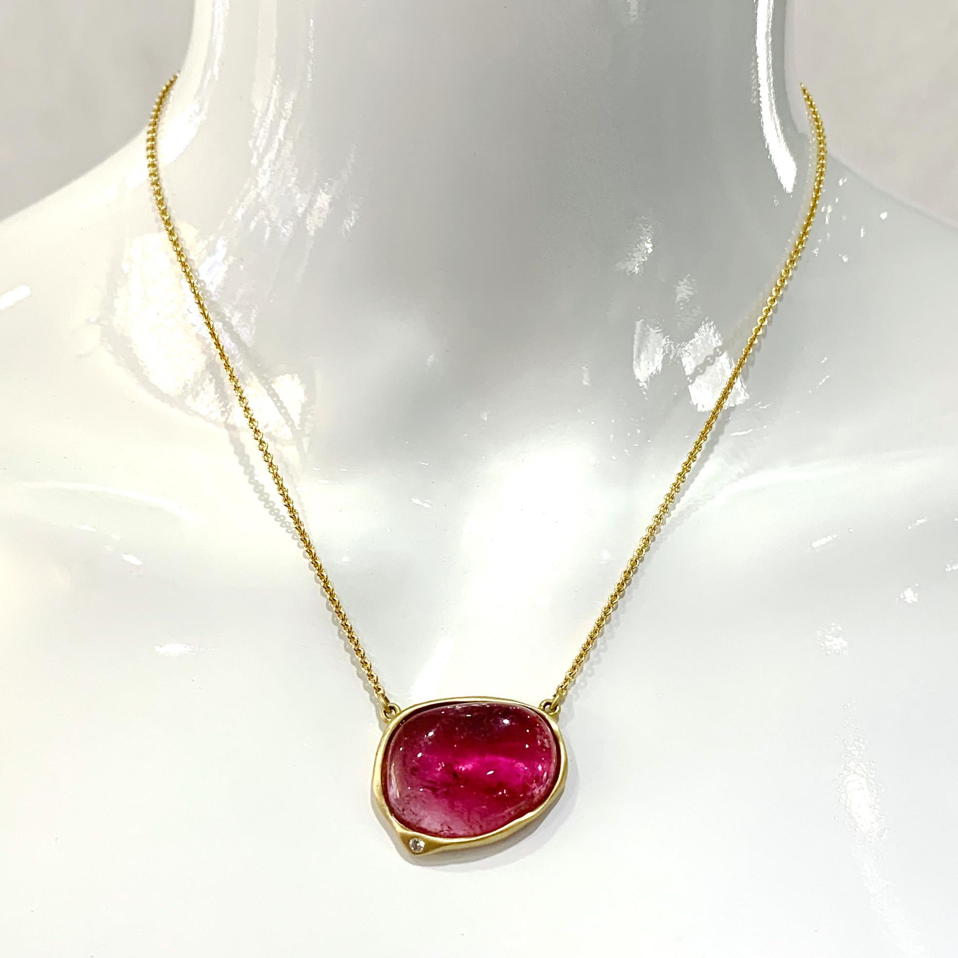 A large roughly egg-shaped cabochon of pink tourmaline sits in a freeform frame of 18 karat yellow gold with a smooth satin finish and a small diamond accent.  Eytan Brandes designed, sculpted and finished this pendant by hand here in our shop.  It